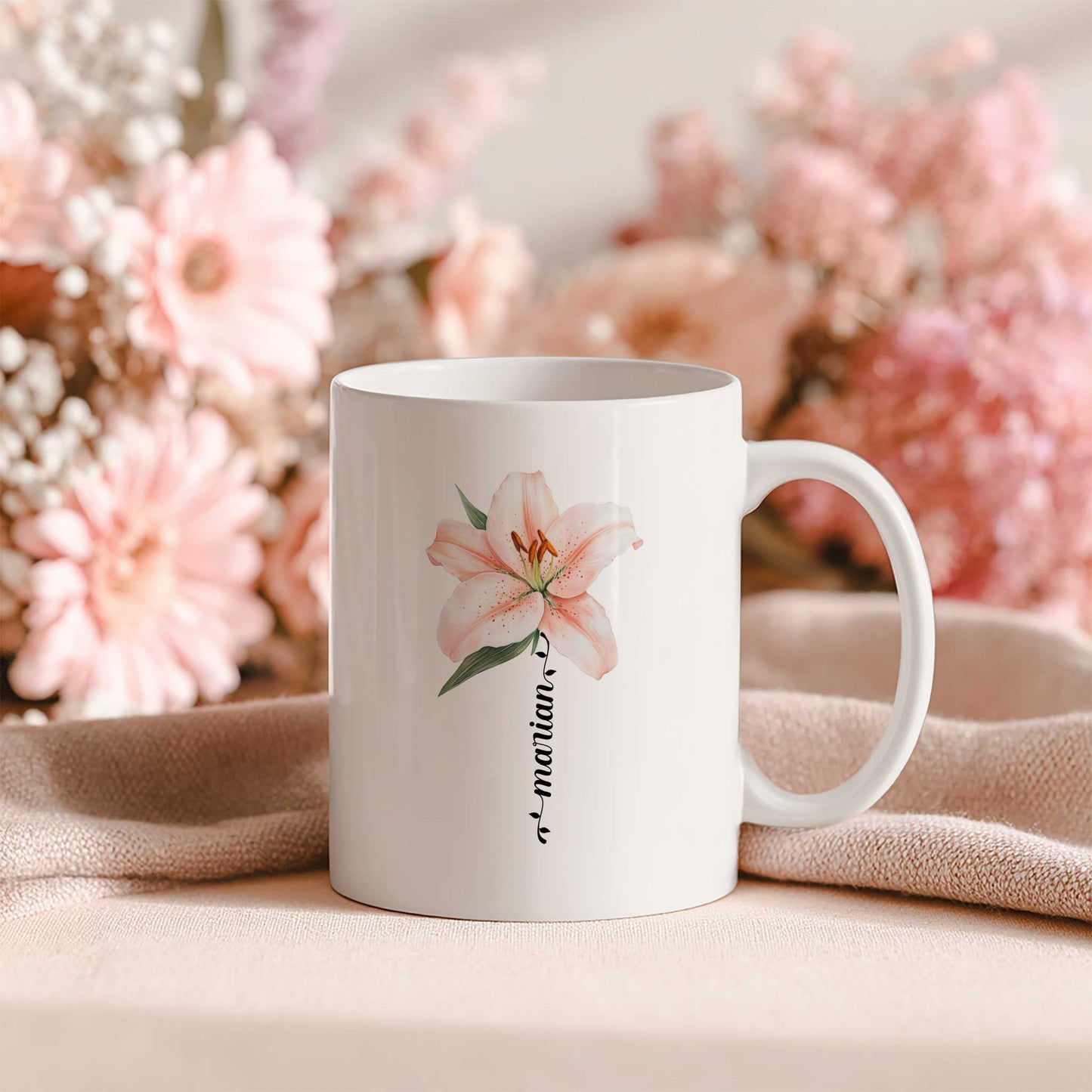 Witte keramische mok met een lichtroze lelie en gepersonaliseerde naam als bloemsteel, geplaatst op een zachte beige stof met roze bloemen op de achtergrond.