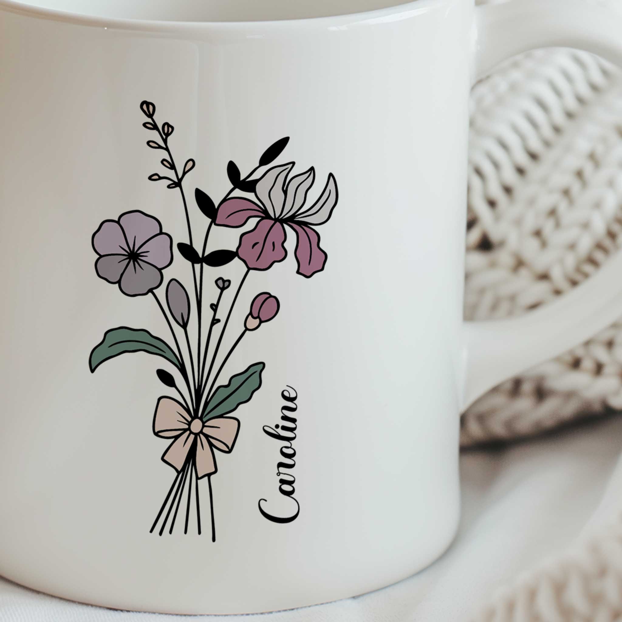 Detailfoto van een witte keramische mok met een boeket lijnbloemen in zachte paars-roze tinten, afgewerkt met een strik. De gepersonaliseerde naam Caroline staat naast het bloemendesign.