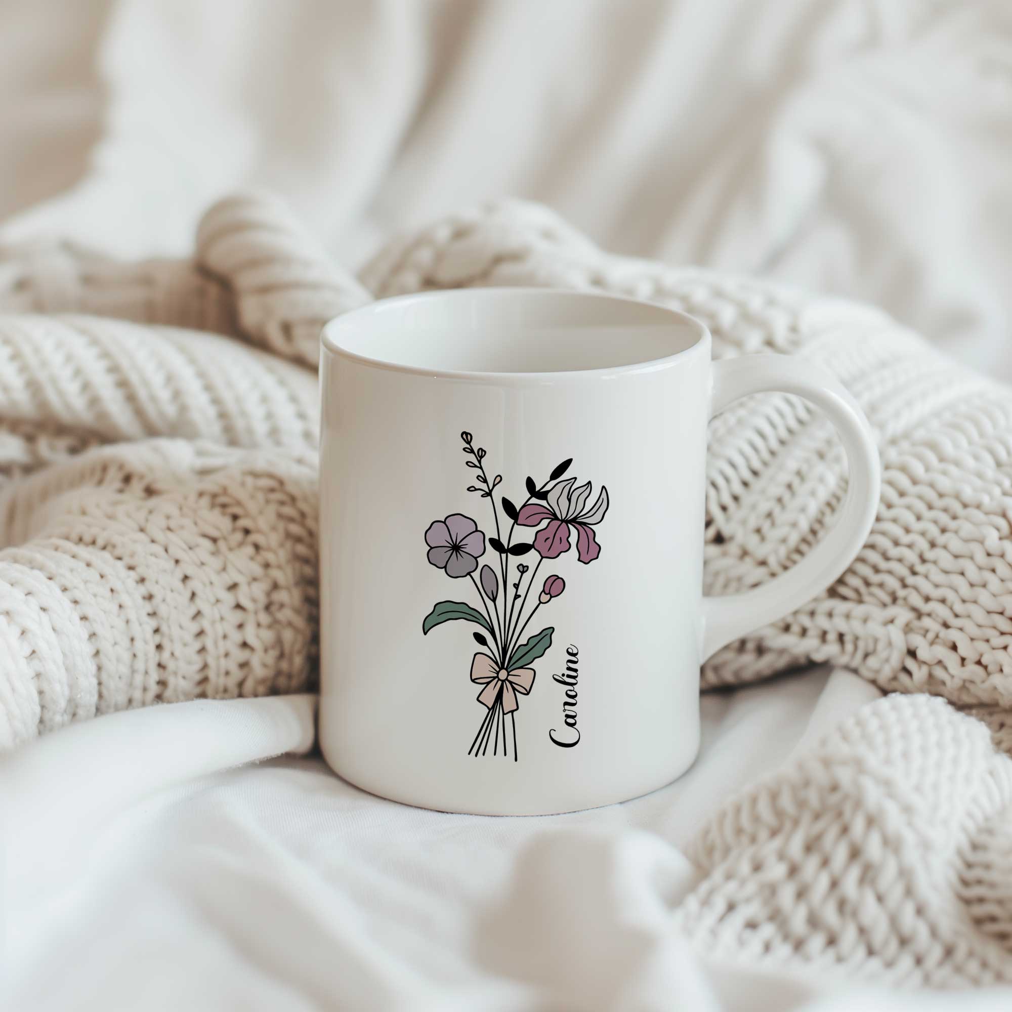 Witte keramische mok met een boeket lijnbloemen in zachte paars-roze tinten en een gepersonaliseerde naam naast het bloemendesign. De mok staat tussen zachte gebreide dekens in crèmekleurige tinten.
