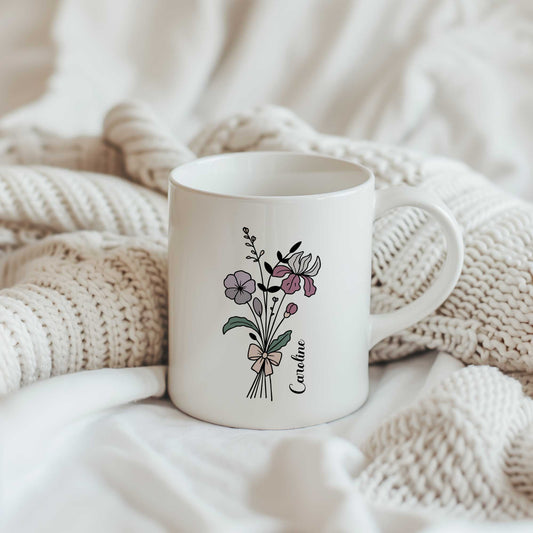 Witte keramische mok met een boeket lijnbloemen in zachte paars-roze tinten en een gepersonaliseerde naam naast het bloemendesign. De mok staat tussen zachte gebreide dekens in crèmekleurige tinten.