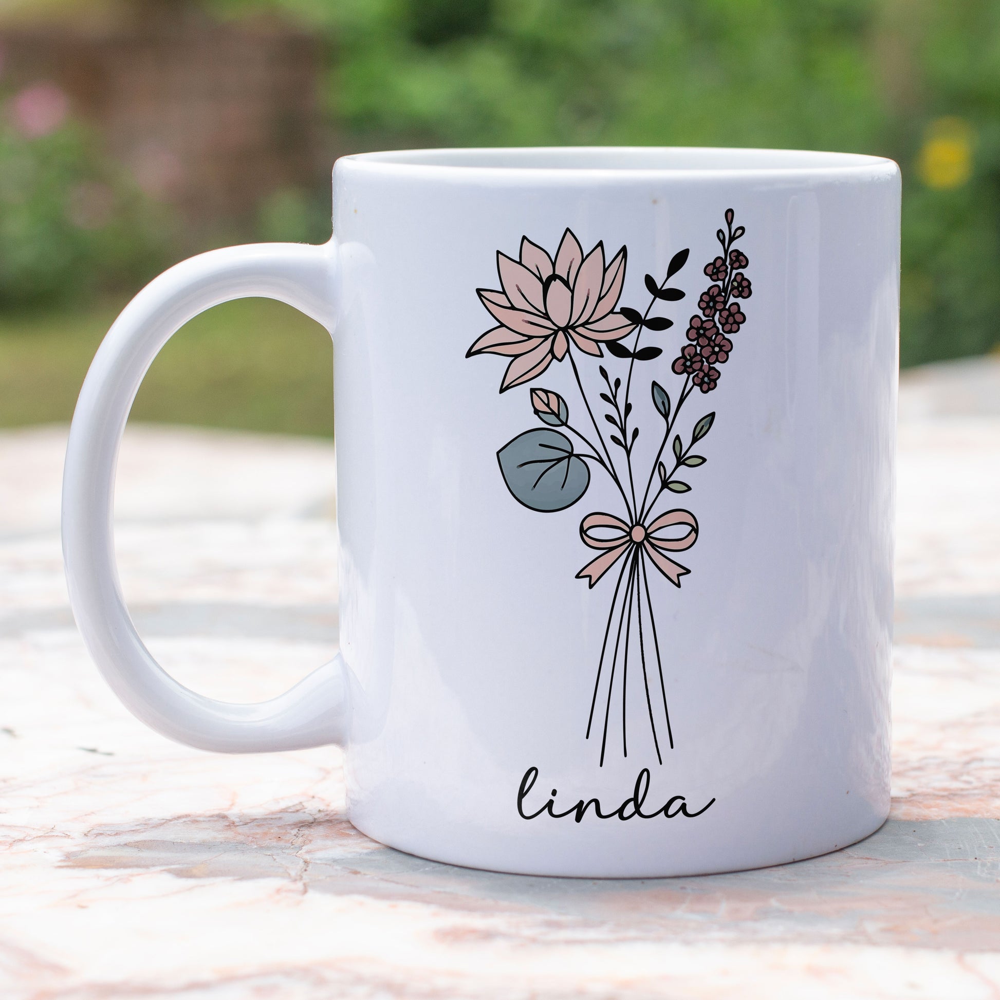 Witte mok met bloemenillustratie en naam Linda, keramiek, geplaatst op buitentafel met groene achtergrond