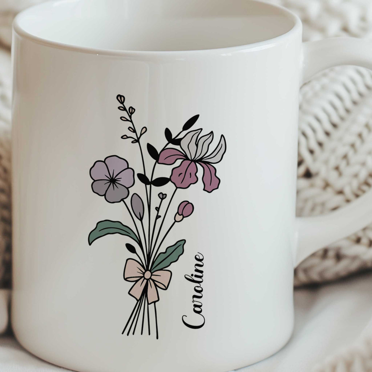 Witte keramische mok met een gepersonaliseerd bloemenboeket en de naam Caroline, omringd door een zachte crèmekleurige gebreide deken die zorgt voor een warme en rustige uitstraling.