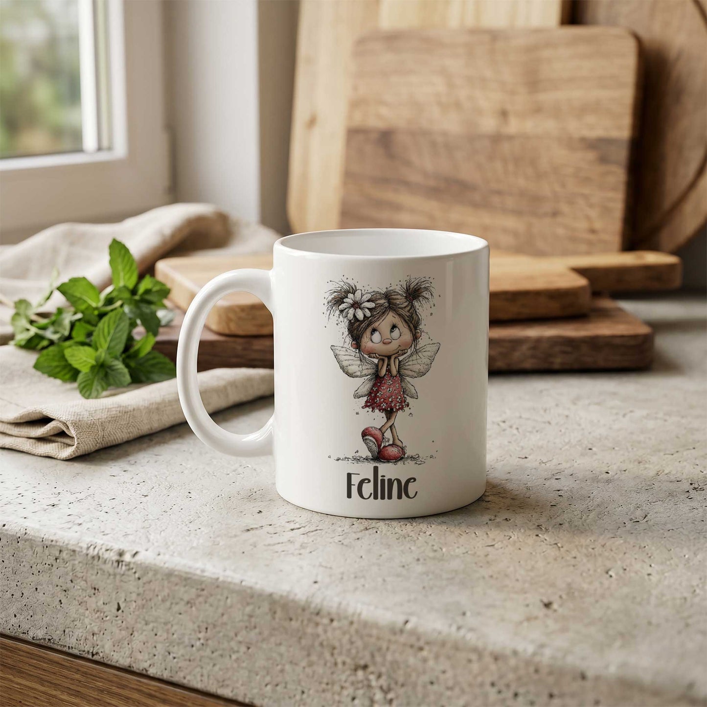 Witte mok met fee-illustratie en naam Feline op een stenen keukenblad met houten snijplanken op de achtergrond.