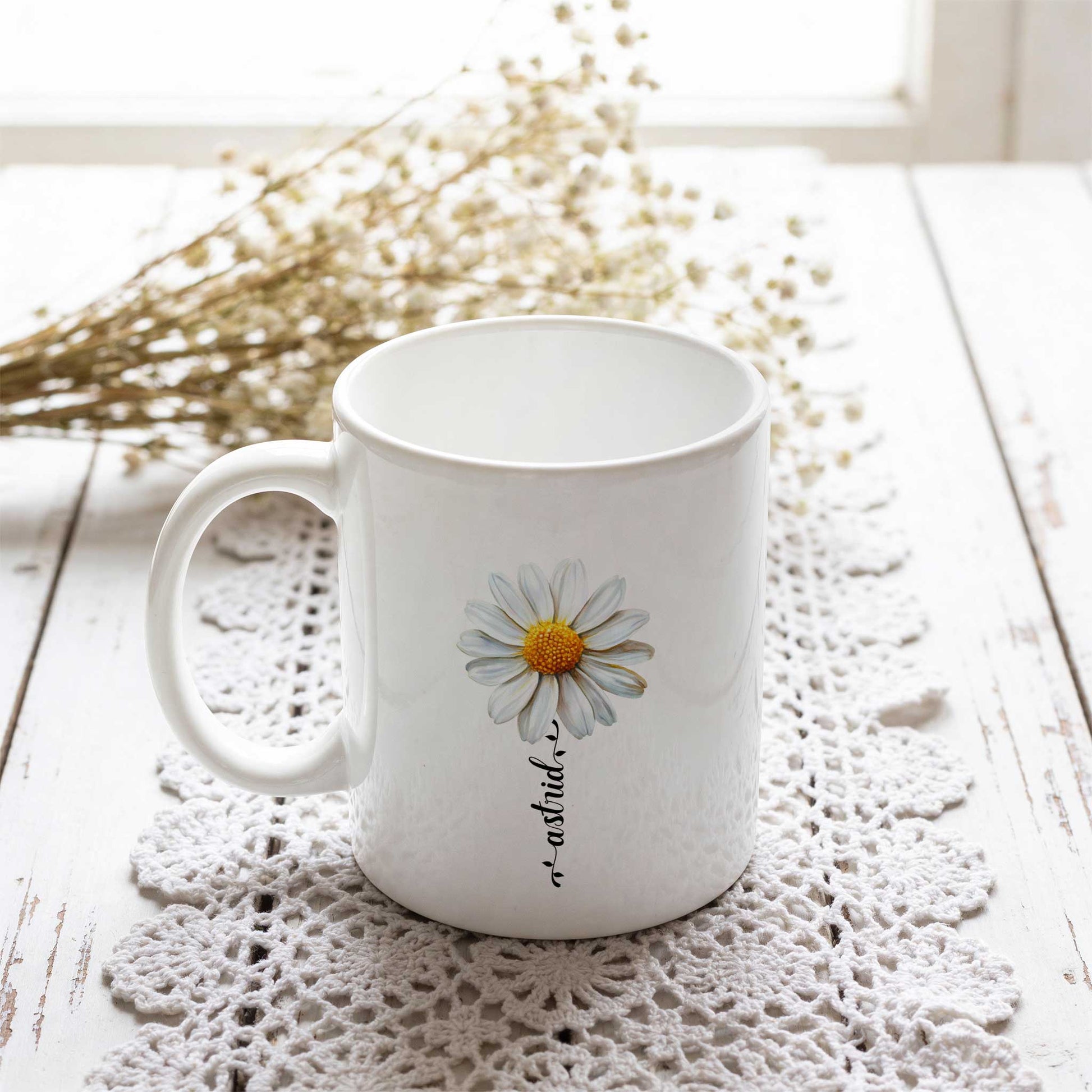 Witte keramische mok met een madeliefbloem en een gepersonaliseerde naam verwerkt in de steel van de bloem. De mok staat op een wit gehaakt kleedje op een lichte houten tafel, met gedroogde bloemetjes op de achtergrond.