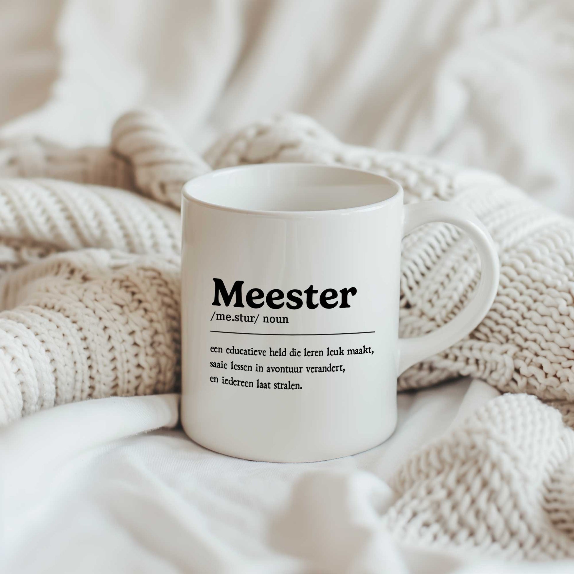 Witte keramische mok bedrukt met de tekst "Meester" en een woordenboekstijl definitie. De mok staat tussen zachte crèmekleurige gebreide dekens.