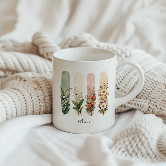 Witte mok met een illustratie van de geboortebloemen van mei en de tekst Mei, geplaatst tussen beige gebreide dekens.