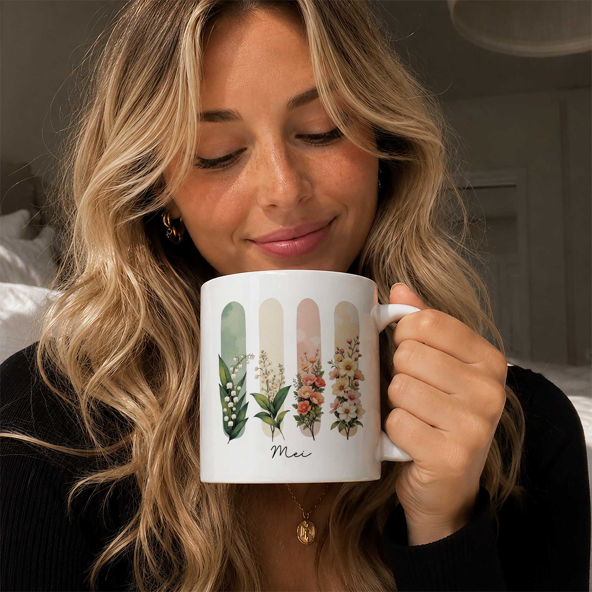 Blonde vrouw houdt een witte mok vast met een illustratie van de geboortebloemen van mei en de tekst Mei.
