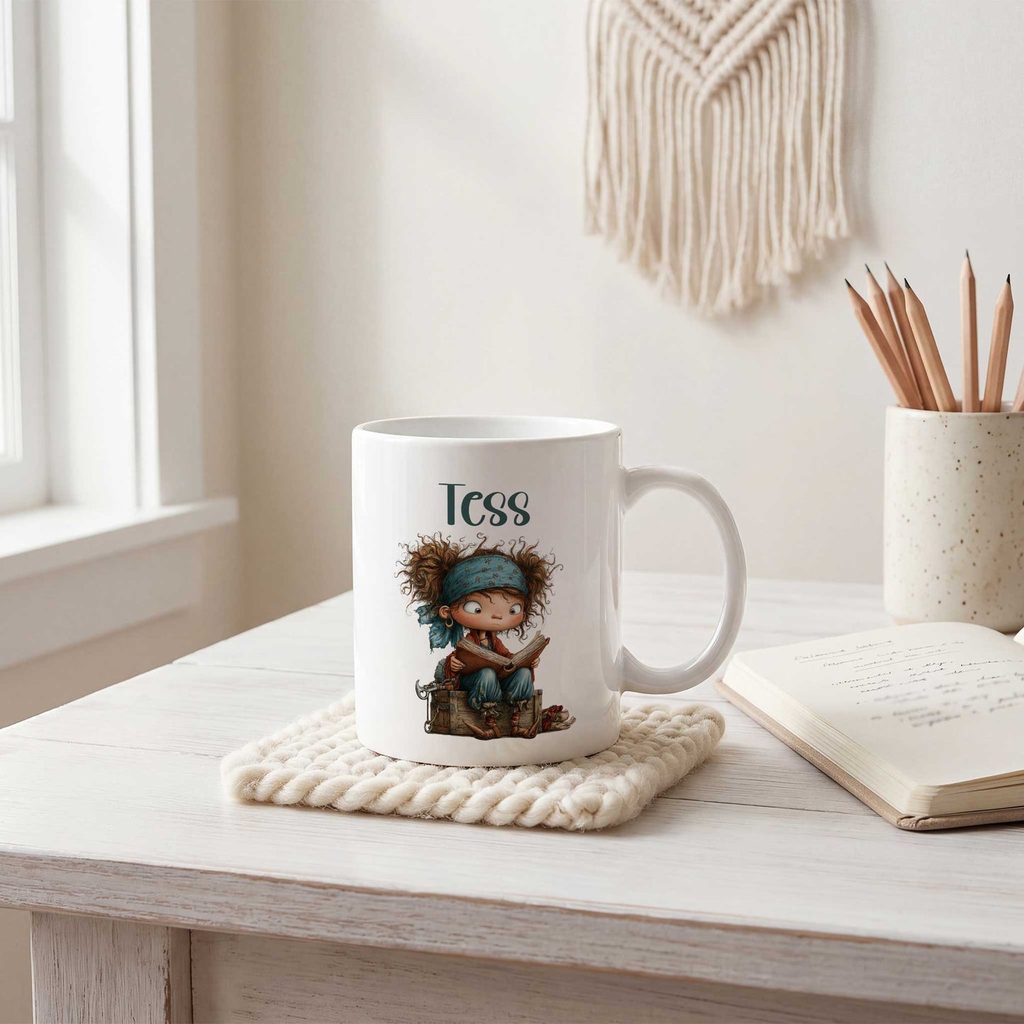 Witte mok op een licht bureau met de naam Tess boven een illustratie van een klein piratenkind dat op een houten kist zit en een boek leest. Naast de mok liggen een notitieboek en een pot met potloden.