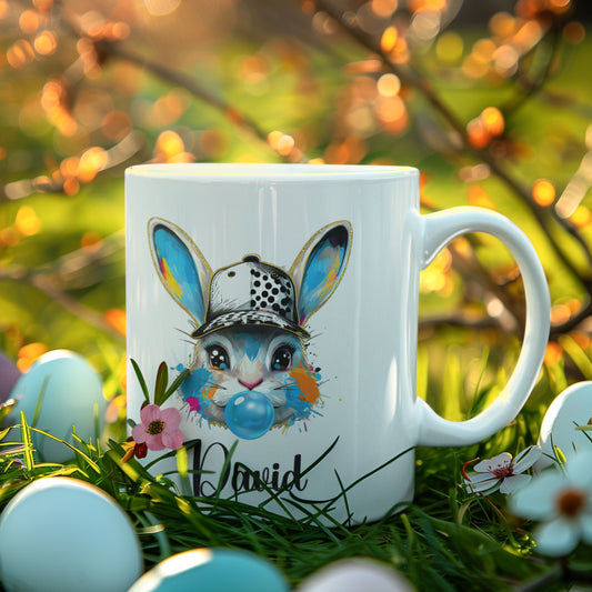 Witte mok met jongenskonijn met kauwgombel, pet met stippen en naam David, omringd door pastelgekleurde paaseieren en gras in een buitenomgeving