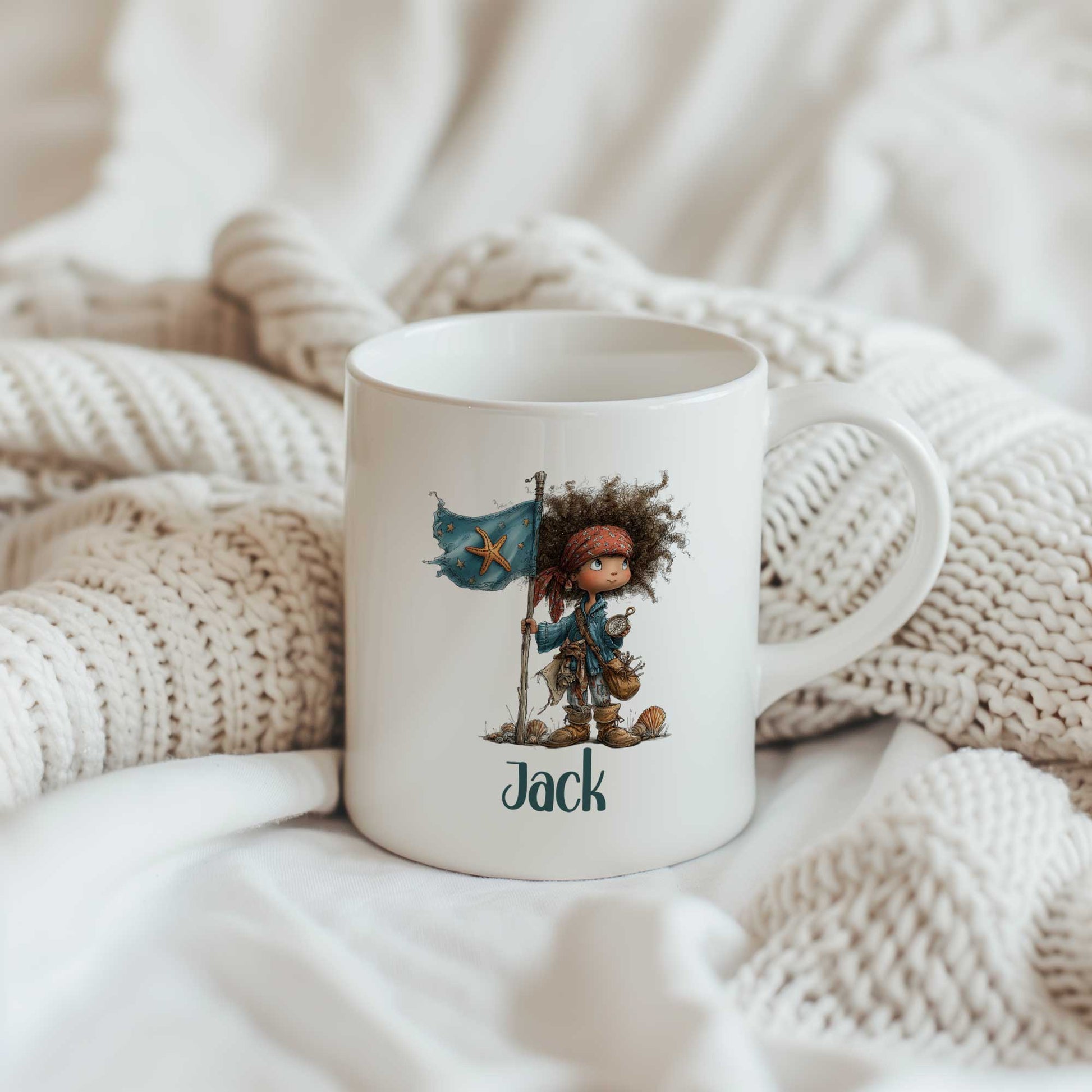 Witte mok met de naam Jack onder een illustratie van een klein piratenkind naast een blauwe vlag met een zeester. De mok staat tussen gebreide dekens.