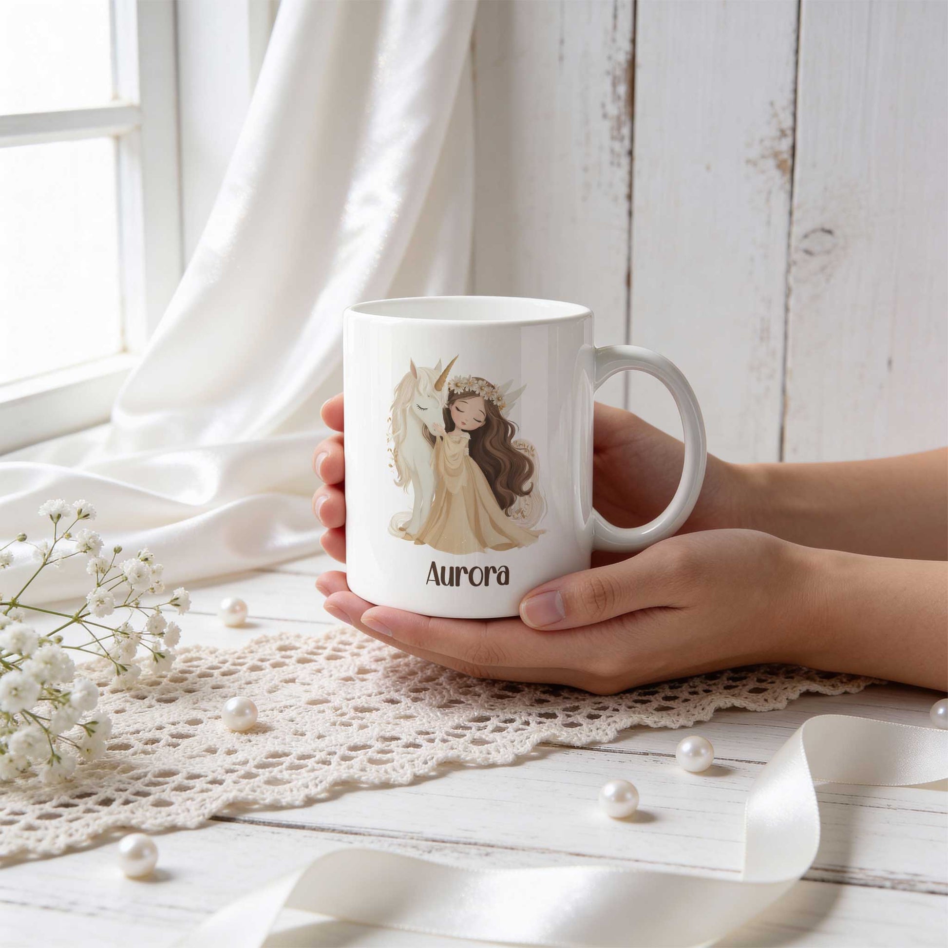 Witte mok met een illustratie van een prinses met lang bruin haar die knuffelt met een witte eenhoorn. Onder de illustratie staat de naam Aurora. De mok wordt vastgehouden boven een gehaakt kleed met witte bloemen en parels.