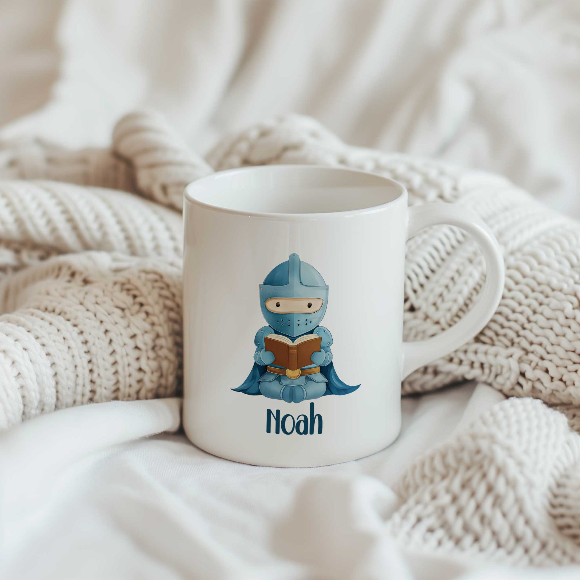 Sfeerfoto van een witte mok met een blauwe ridder die een boek leest en de naam Noah op een gebreide deken.