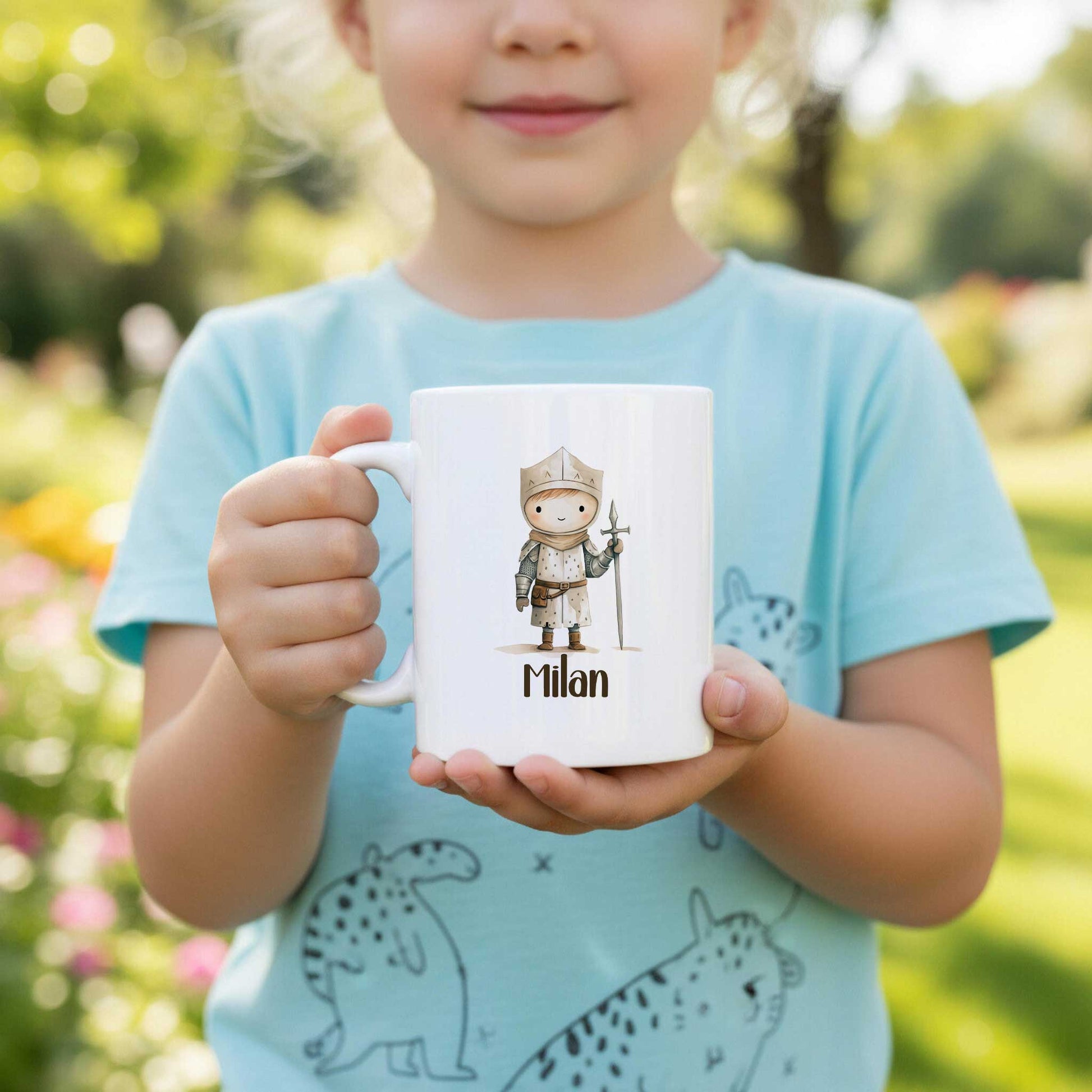 Kind houdt buiten een witte mok vast met een illustratie van een kleine ridder met kroon, een zwaard en de naam Milan. Op de achtergrond zijn bomen en planten te zien.
