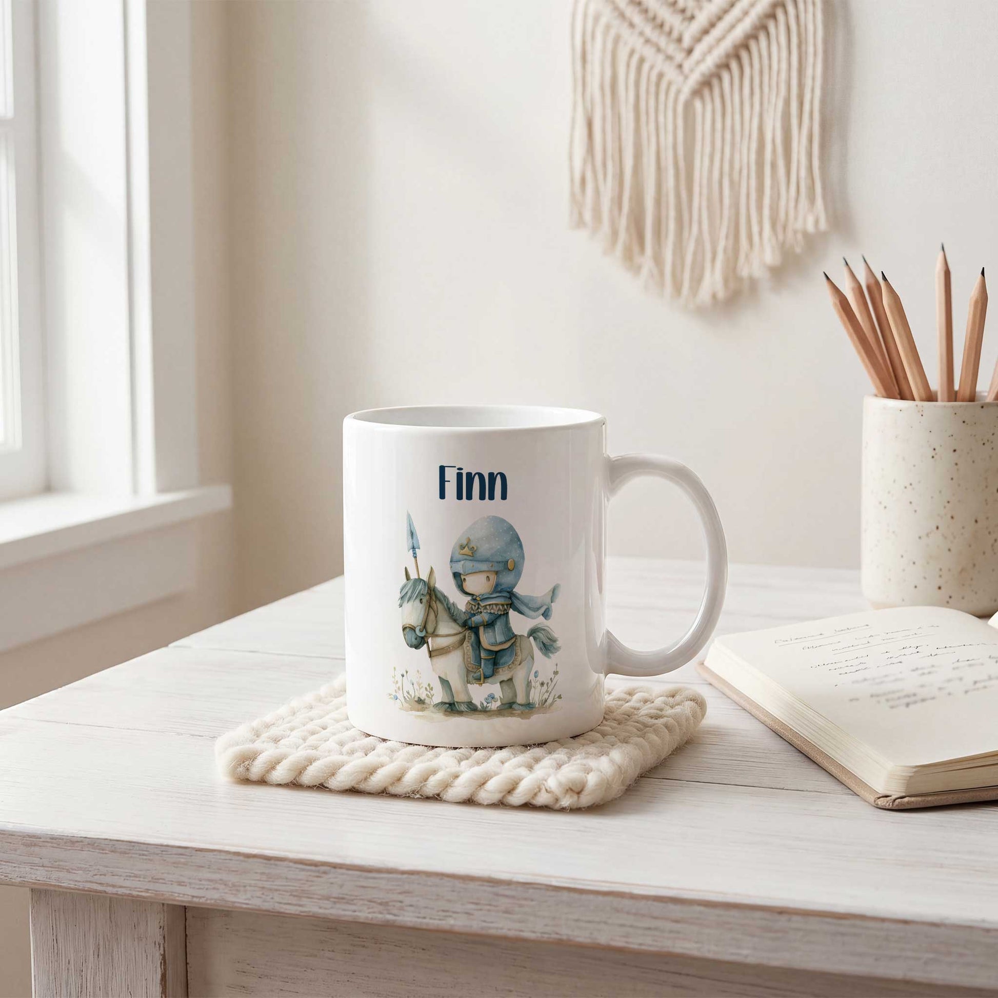 Witte keramische mok met de naam Finn en een illustratie van een lieve blauwe ridder op een wit paard, geplaatst op een witte tafel naast een notitieboek, potloodhouder en raam.