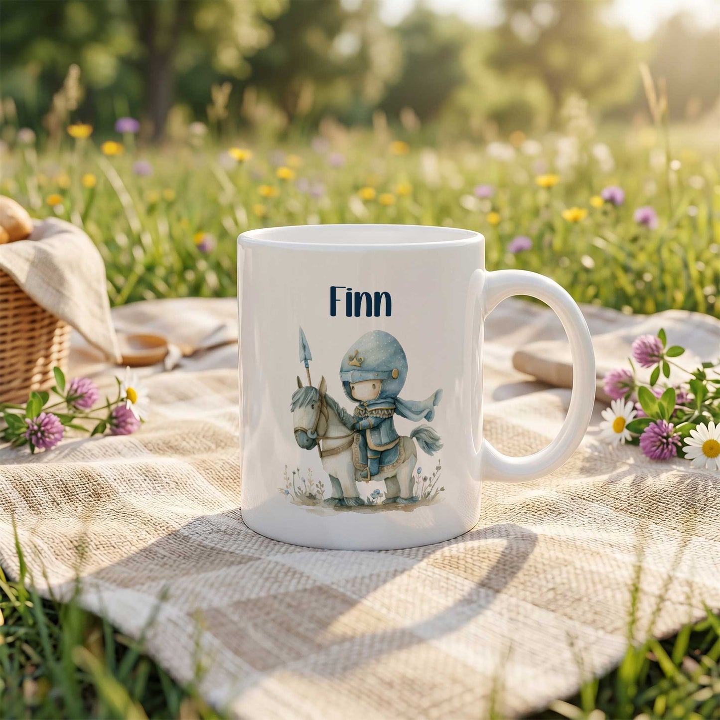 Witte keramische mok met de naam Finn en een illustratie van een lieve blauwe ridder op een wit paard, geplaatst op een picknickkleed in een bloemenweide met gras, bloemen en bomen op de achtergrond.