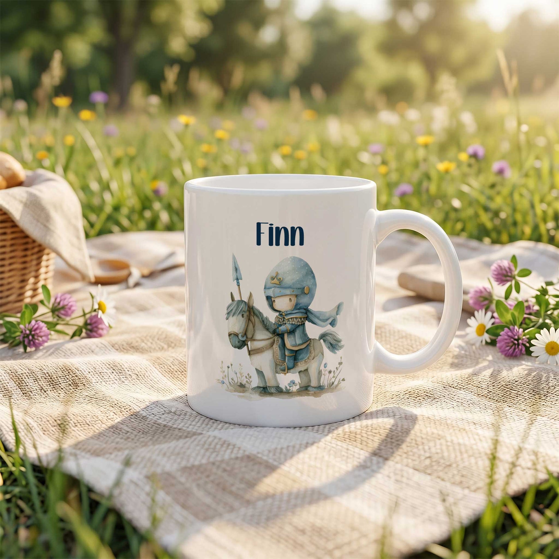 Witte keramische mok met de naam Finn en een illustratie van een lieve blauwe ridder op een wit paard, geplaatst op een picknickkleed in een bloemenweide met gras, bloemen en bomen op de achtergrond.