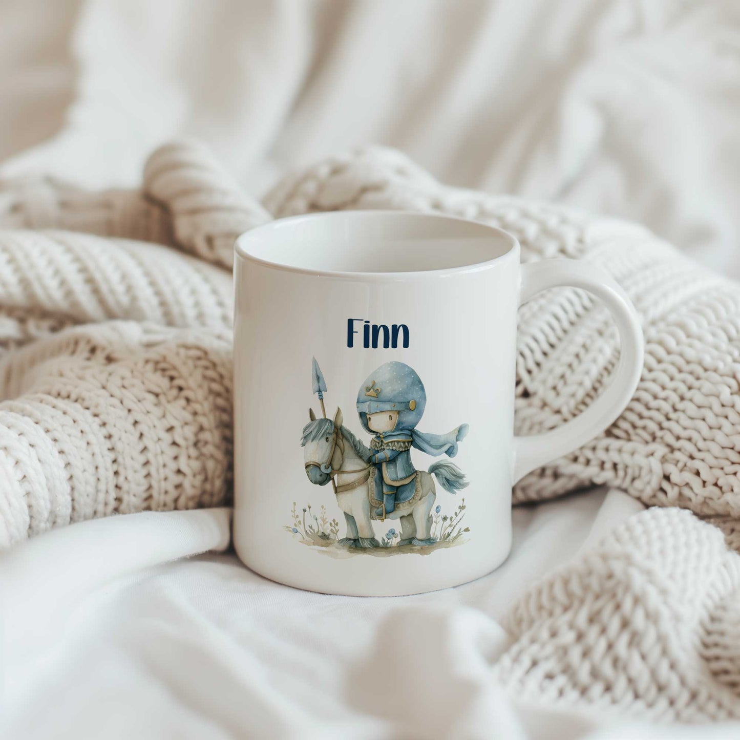 Witte keramische mok met de naam Finn en een illustratie van een lieve blauwe ridder op een wit paard, geplaatst op een gebreide beige deken.