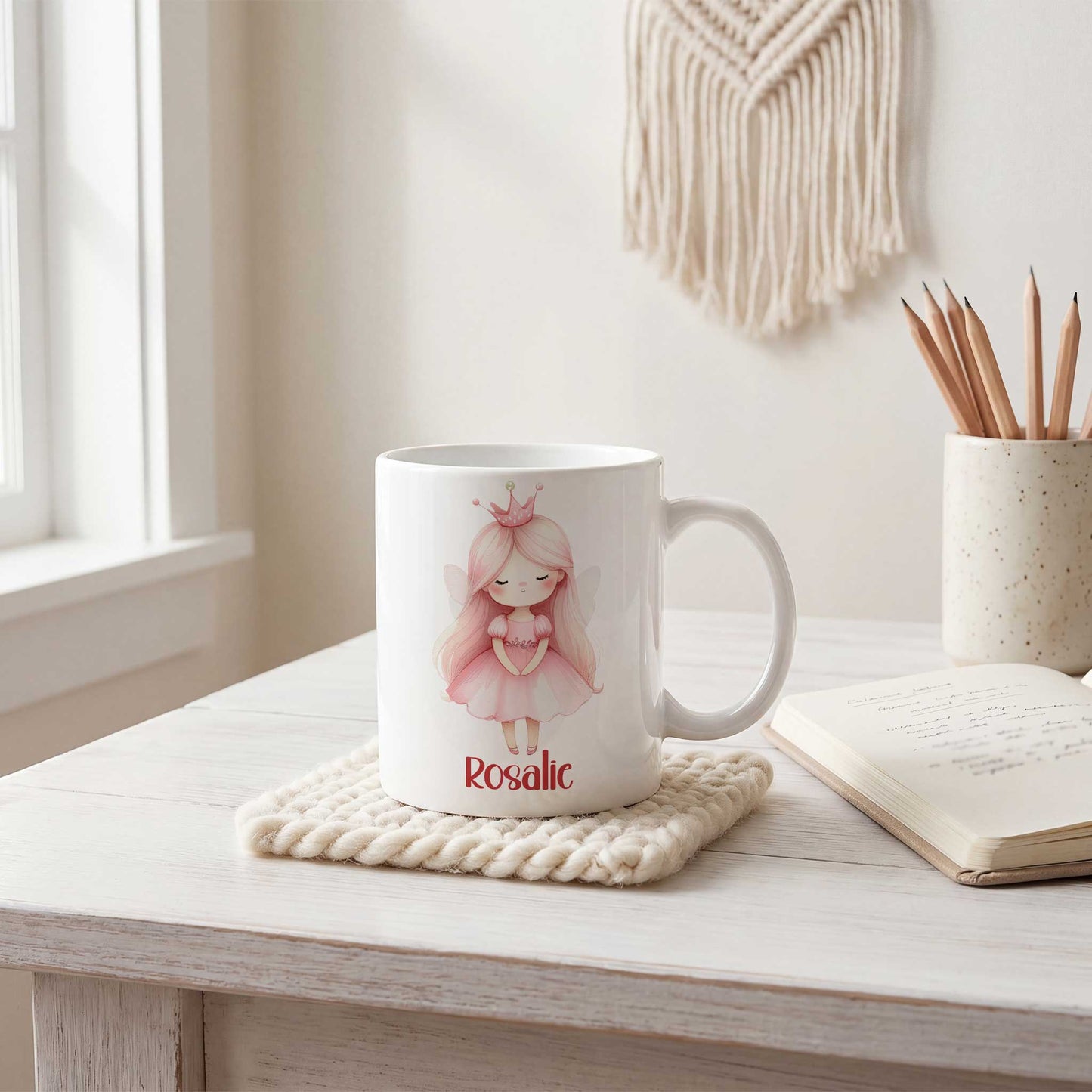 Witte mok met een prinses met roze kroontje en de naam Rosalie op een licht houten bureau naast een notitieboek en een beker met potloden.