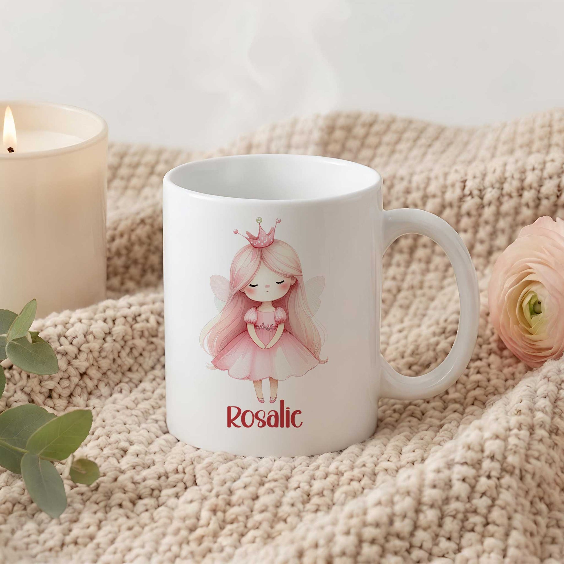 Witte mok met een prinses met roze kroontje en de naam Rosalie op een gebreide beige deken naast een brandende kaars en een lichtroze bloem.
