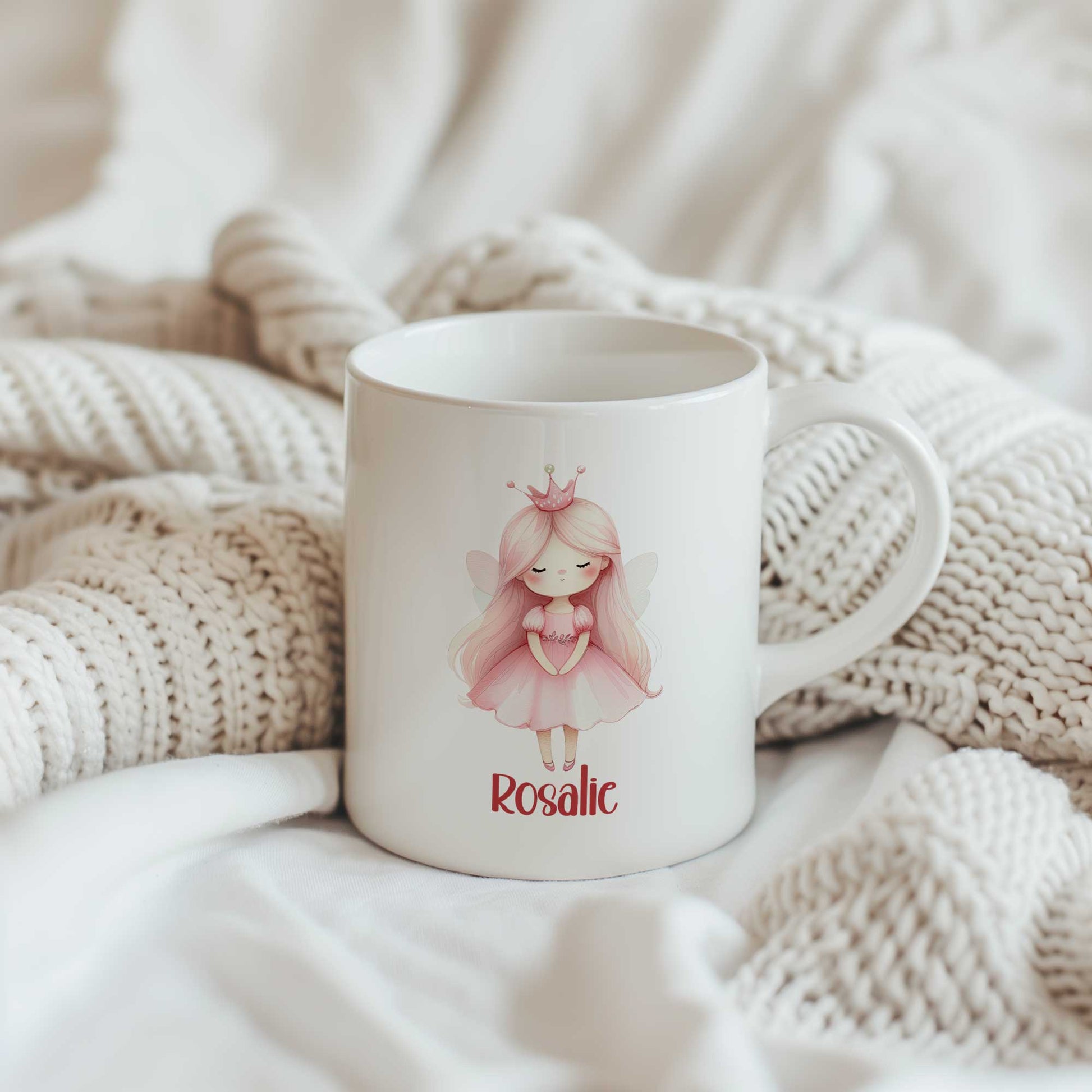 Witte mok met een illustratie van een prinses met lang roze haar, een roze kroontje en een roze jurk. Onder de illustratie staat de naam Rosalie. De mok staat tussen gebreide beige dekens.