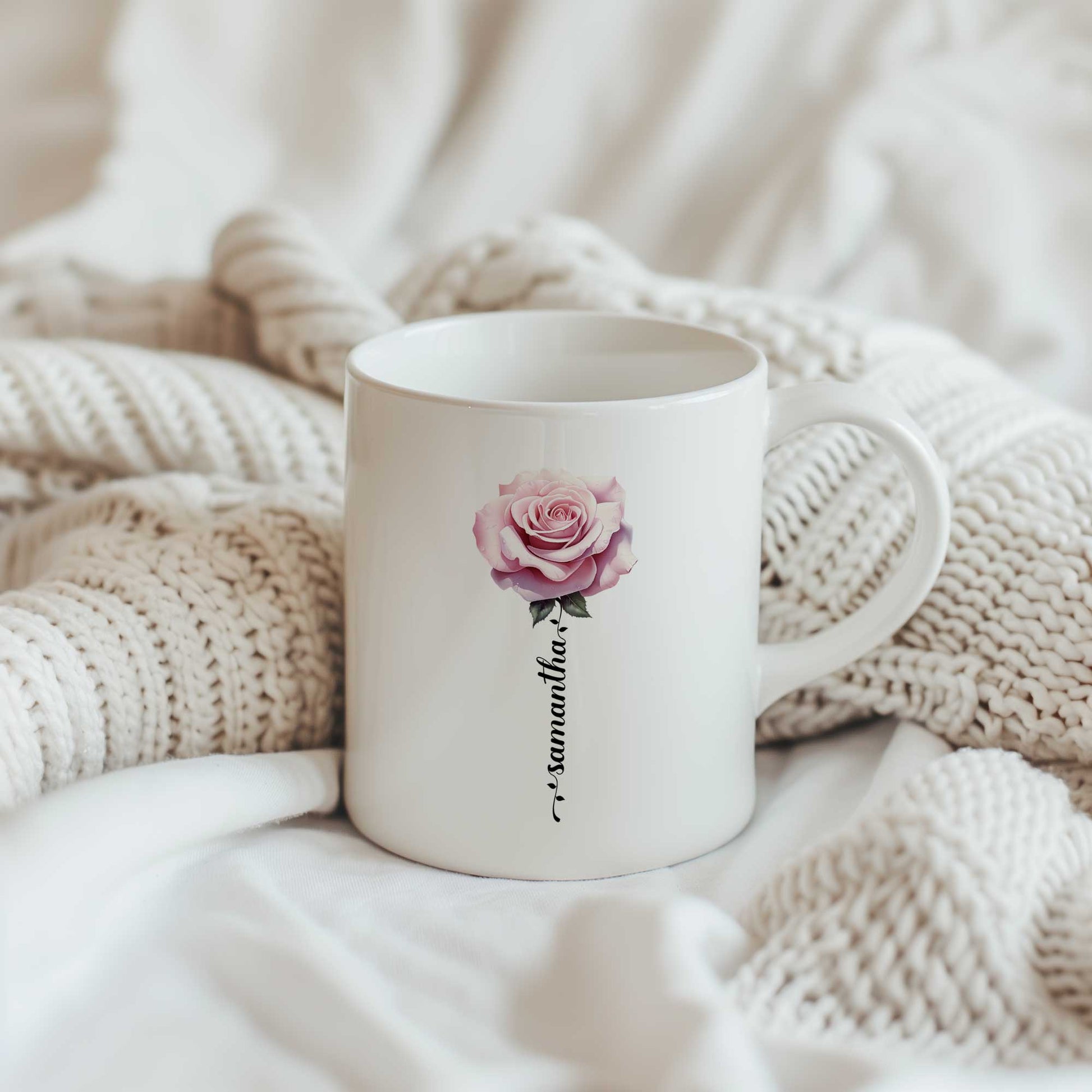 Witte keramische mok met een roze roos en gepersonaliseerde naam als bloemsteel, geplaatst op een zachte beige gebreide deken.