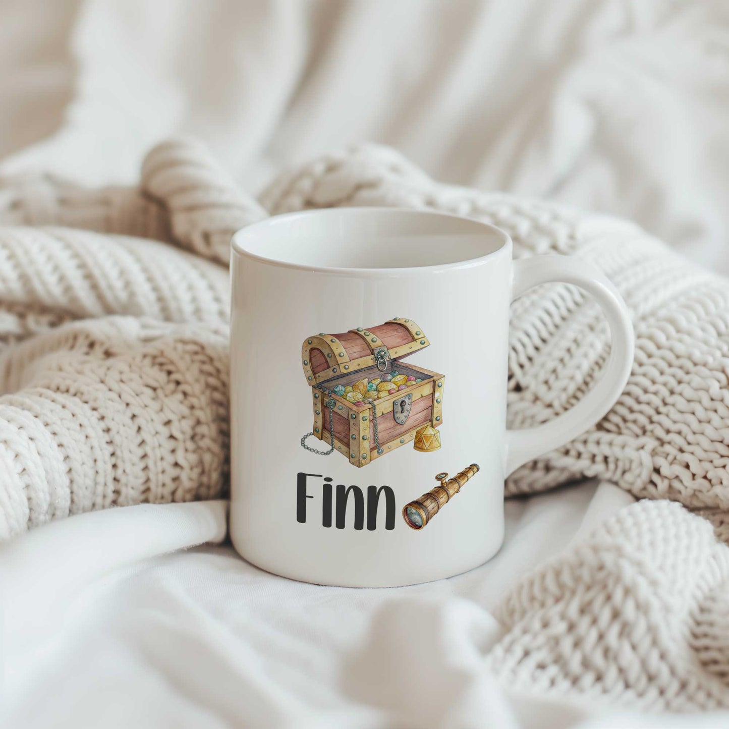 Productfoto van een witte mok met een schatkist, een verrekijker en de naam Finn op een beige gebreide ondergrond.
