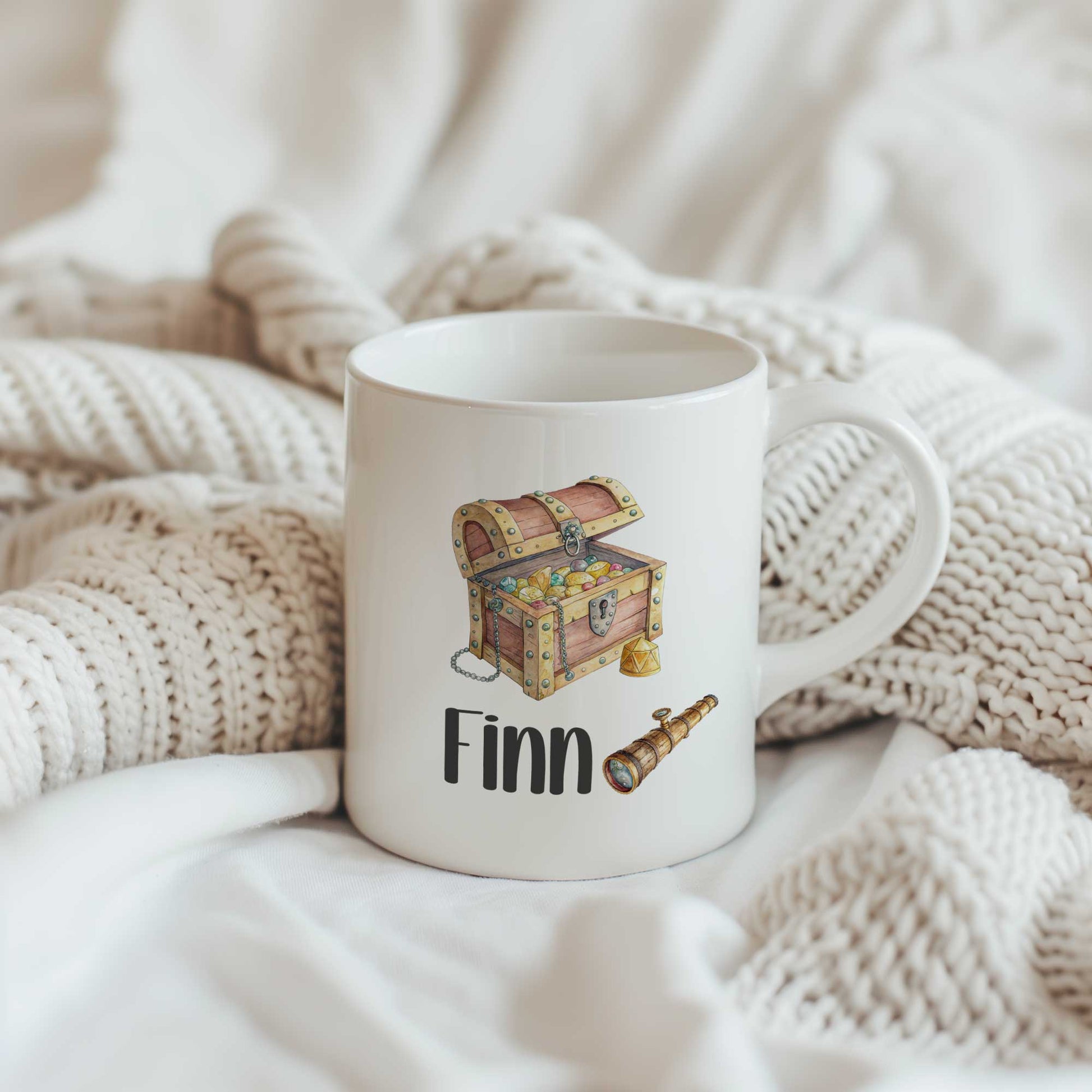 Productfoto van een witte mok met een schatkist, een verrekijker en de naam Finn op een beige gebreide ondergrond.