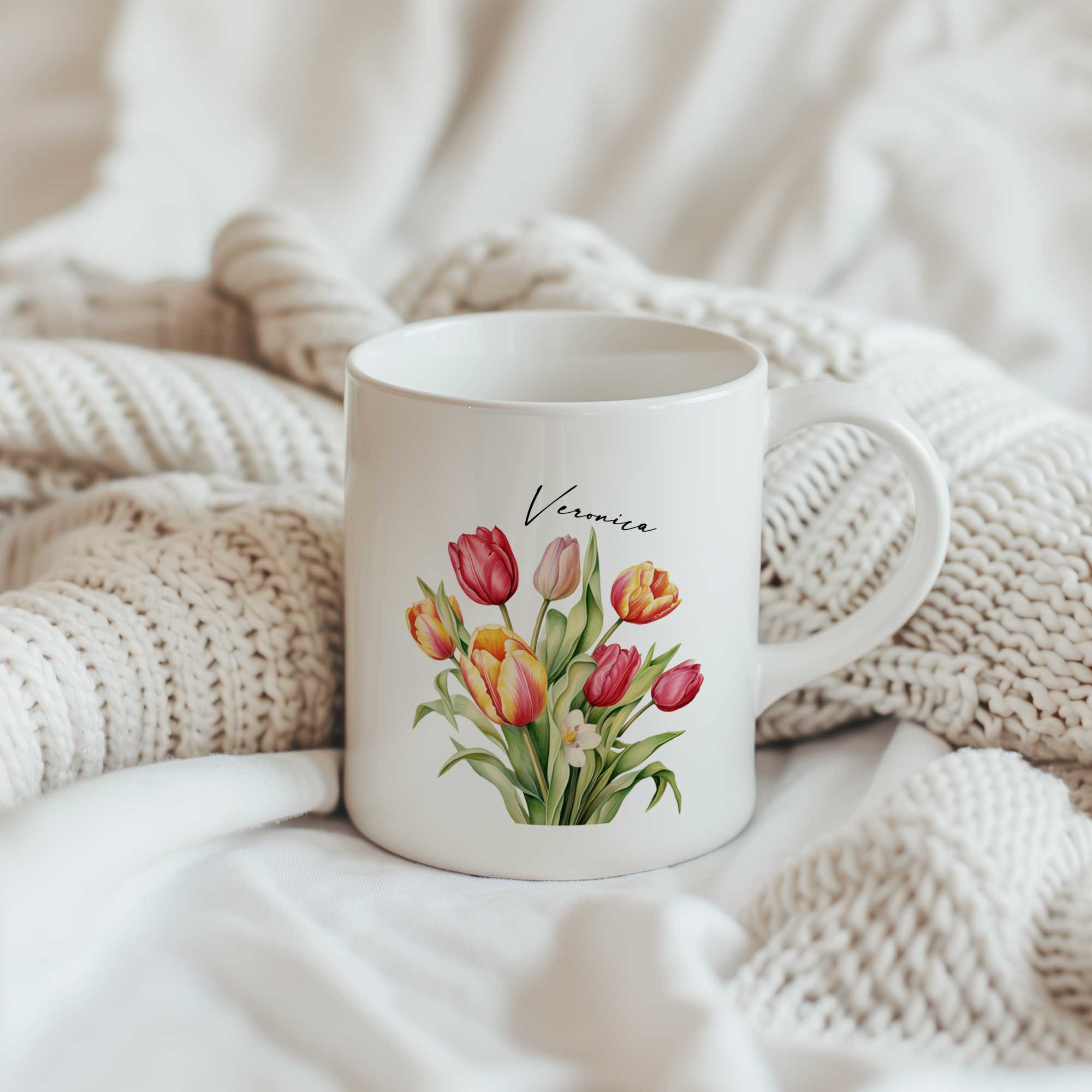 Witte keramische mok met een boeket kleurrijke tulpen en een gepersonaliseerde naam boven de bloemen, geplaatst tussen zachte crèmekleurige gebreide dekens.