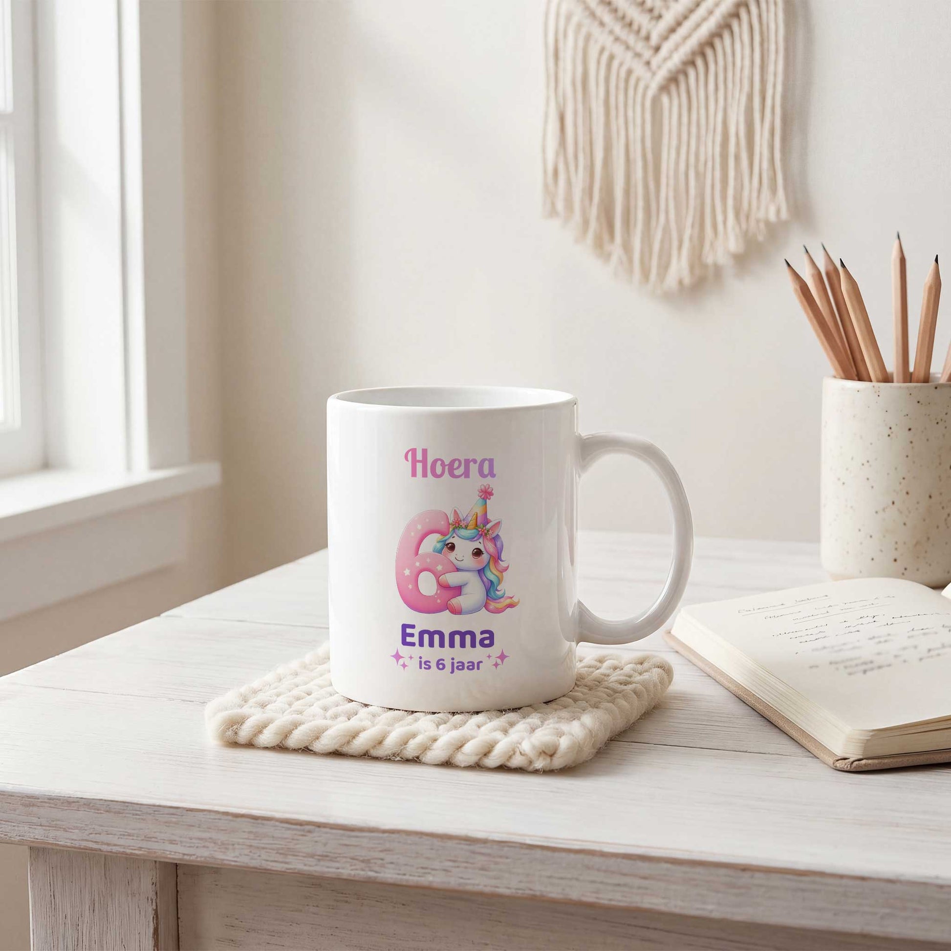 Witte mok met de tekst 'Hoera Emma is 6 jaar' op een gehaakt onderzettertje naast een notitieboek en een pot met potloden.