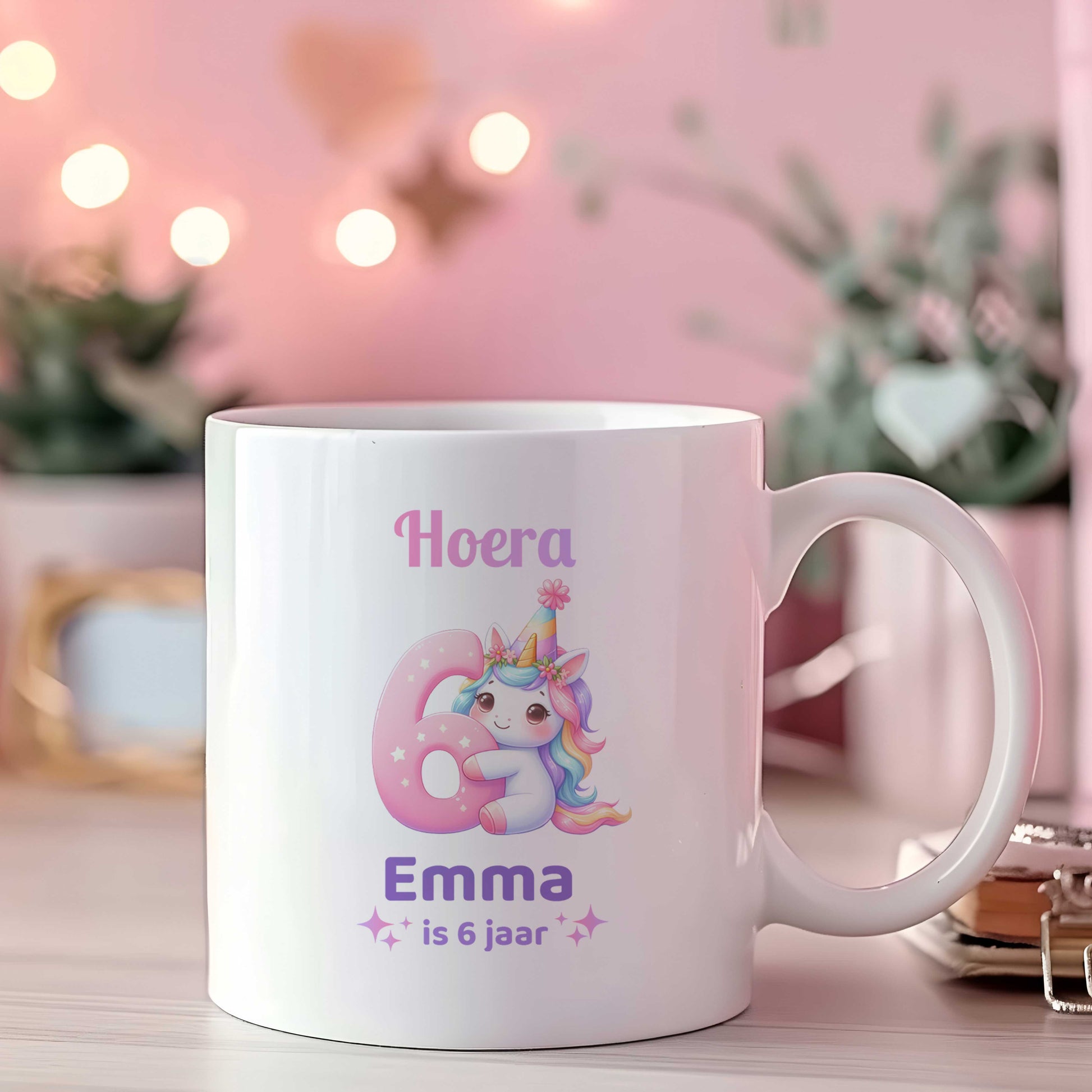 Witte mok op een houten tafel met een unicorn die een groot roze cijfer 6 vasthoudt. Boven de illustratie staat 'Hoera' en onderaan 'Emma is 6 jaar'. Op de achtergrond hangen roze lampjes.