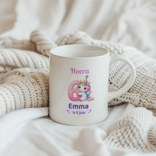 Witte mok met de tekst 'Hoera Emma is 6 jaar' tussen beige gebreide dekens. Op de mok staat een unicorn met een groot roze cijfer 6.