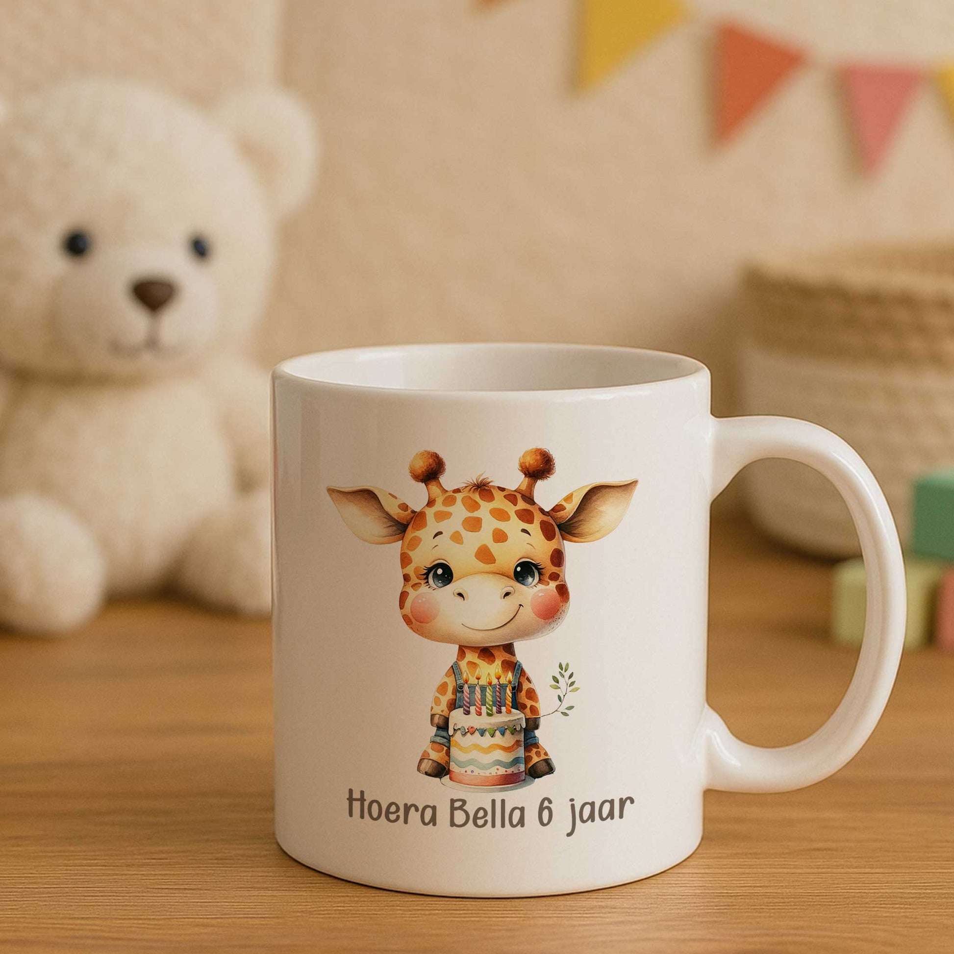 Witte mok met een giraf, verjaardagstaart en de tekst ‘Hoera Bella 6 jaar’ op een houten tafel in een kinderkamer met een grote knuffelbeer en vlaggetjes op de achtergrond.