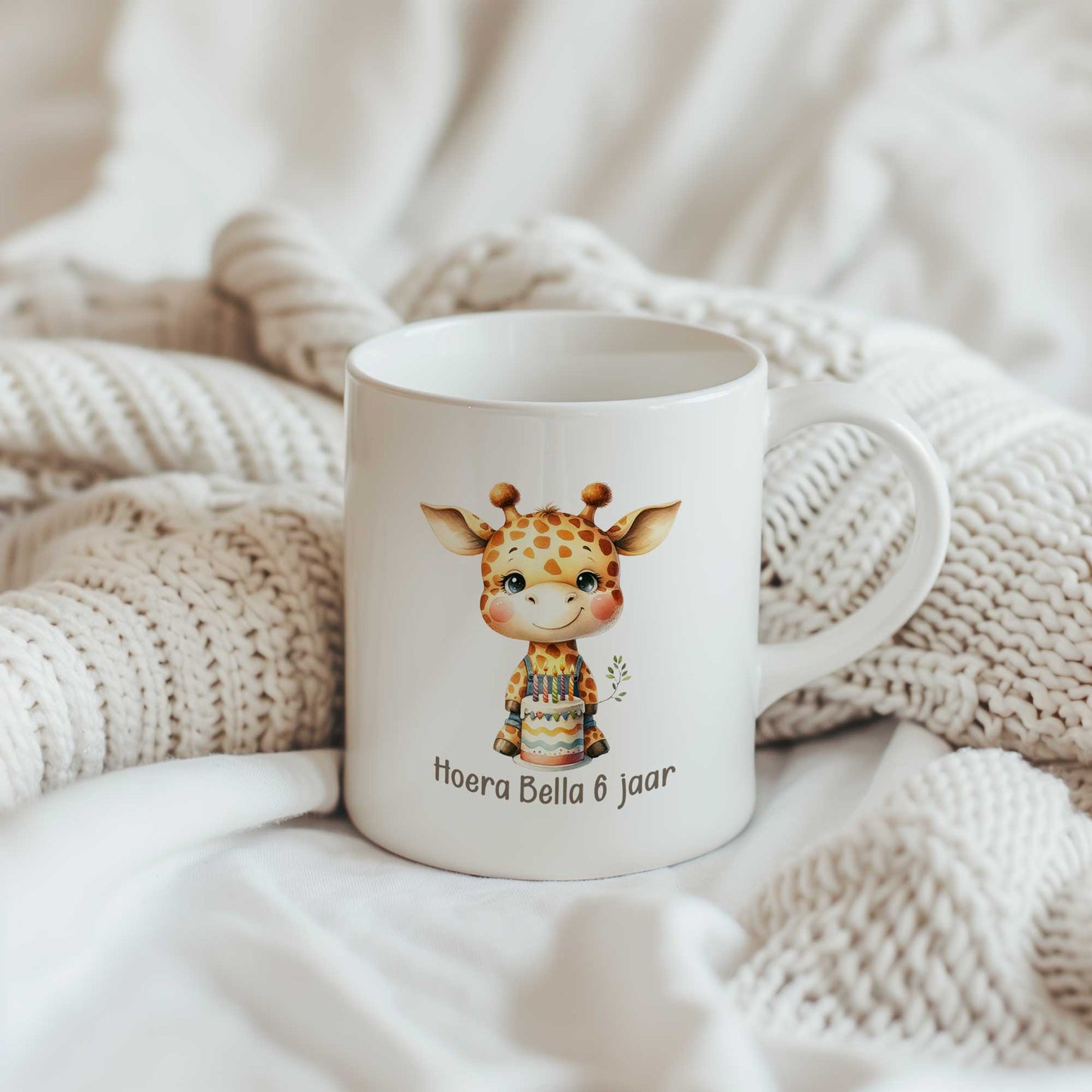 Witte mok met een giraf, verjaardagstaart en de tekst ‘Hoera Bella 6 jaar’ tussen beige gebreide dekens op een bed.
