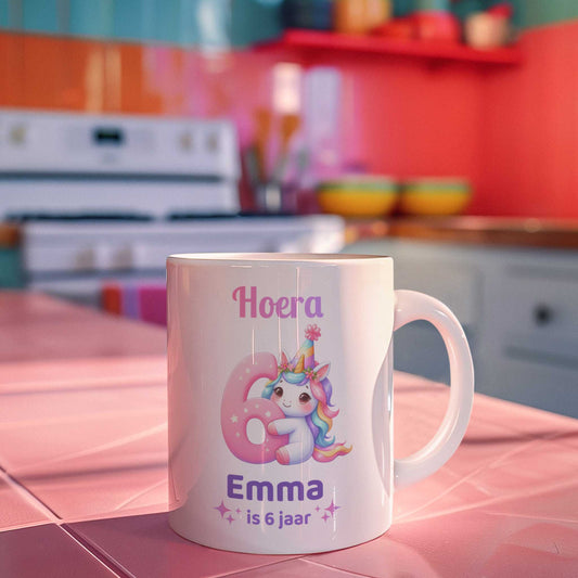 Witte keramische mok met unicorn-illustratie en tekst ‘Hoera Emma is 6 jaar’ op roze keukenachtergrond