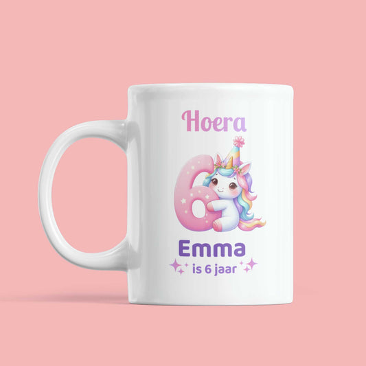 Witte mok met unicorn-illustratie en tekst ‘Hoera Emma is 6 jaar’ tegen roze achtergrond, vooraanzicht