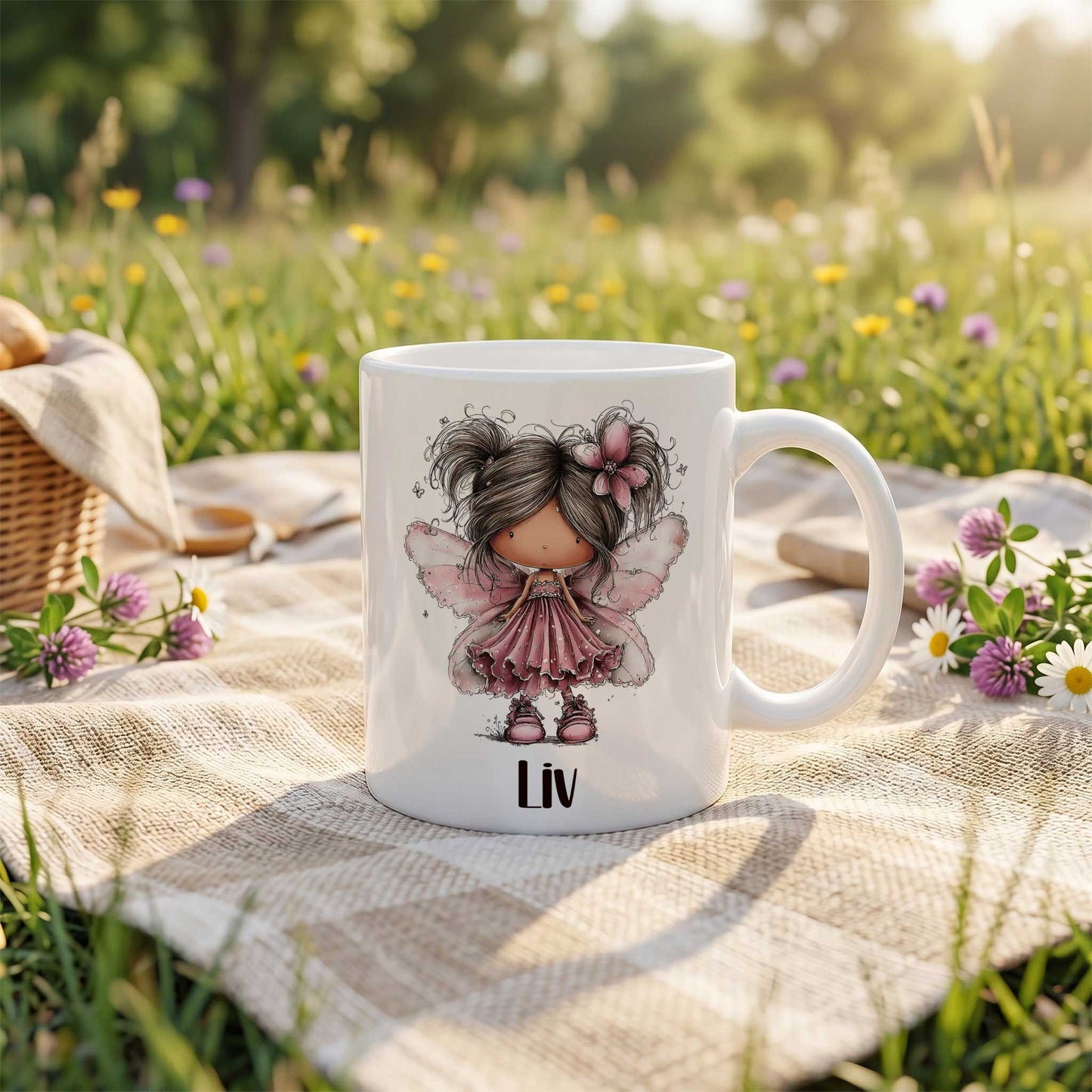 Witte mok met fee-illustratie en naam Liv op een kleed in het gras, met bloemen en een picknickmand op de achtergrond.