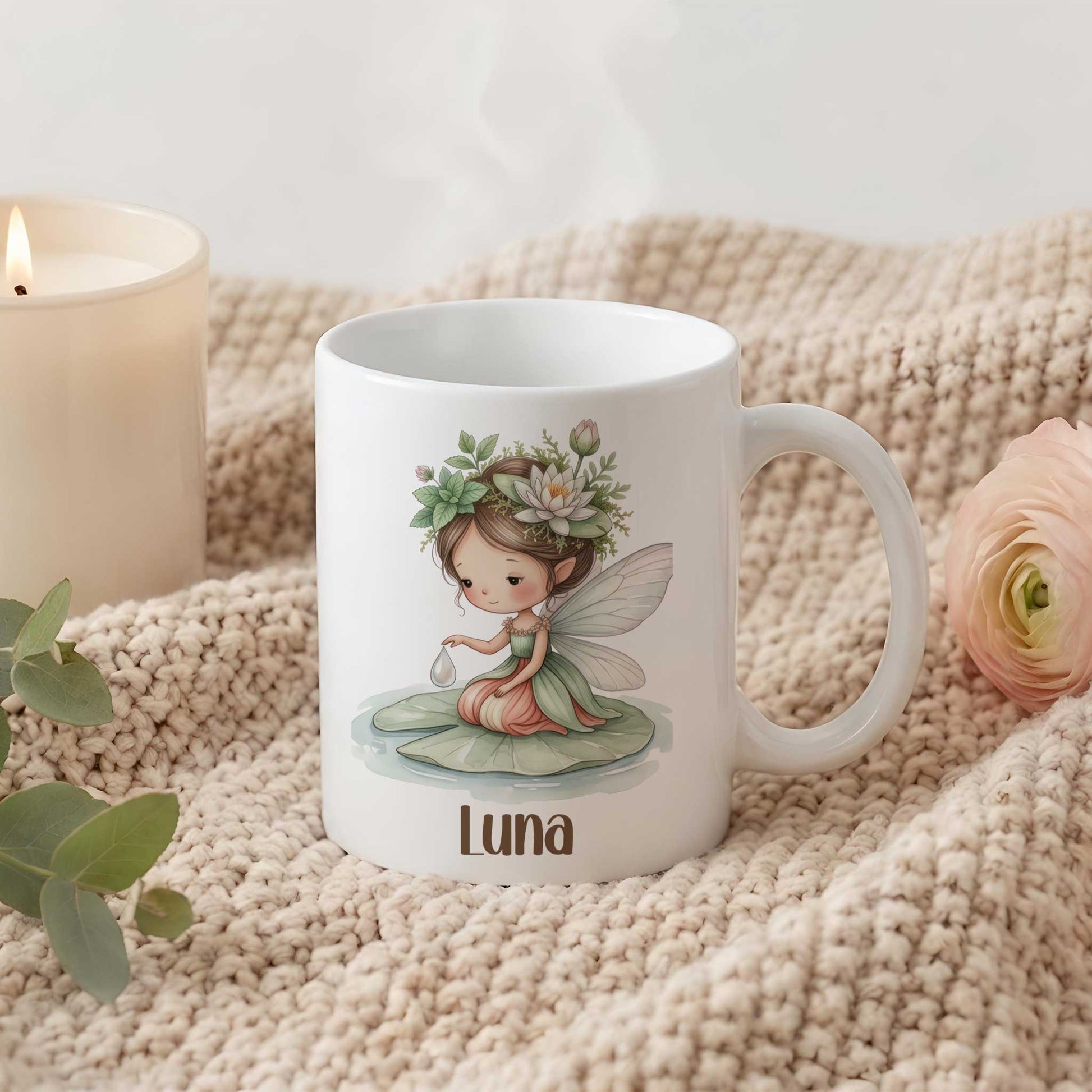 Witte mok met waterleliefee en naam Luna op een beige gebreide deken naast een kaars en bloem.