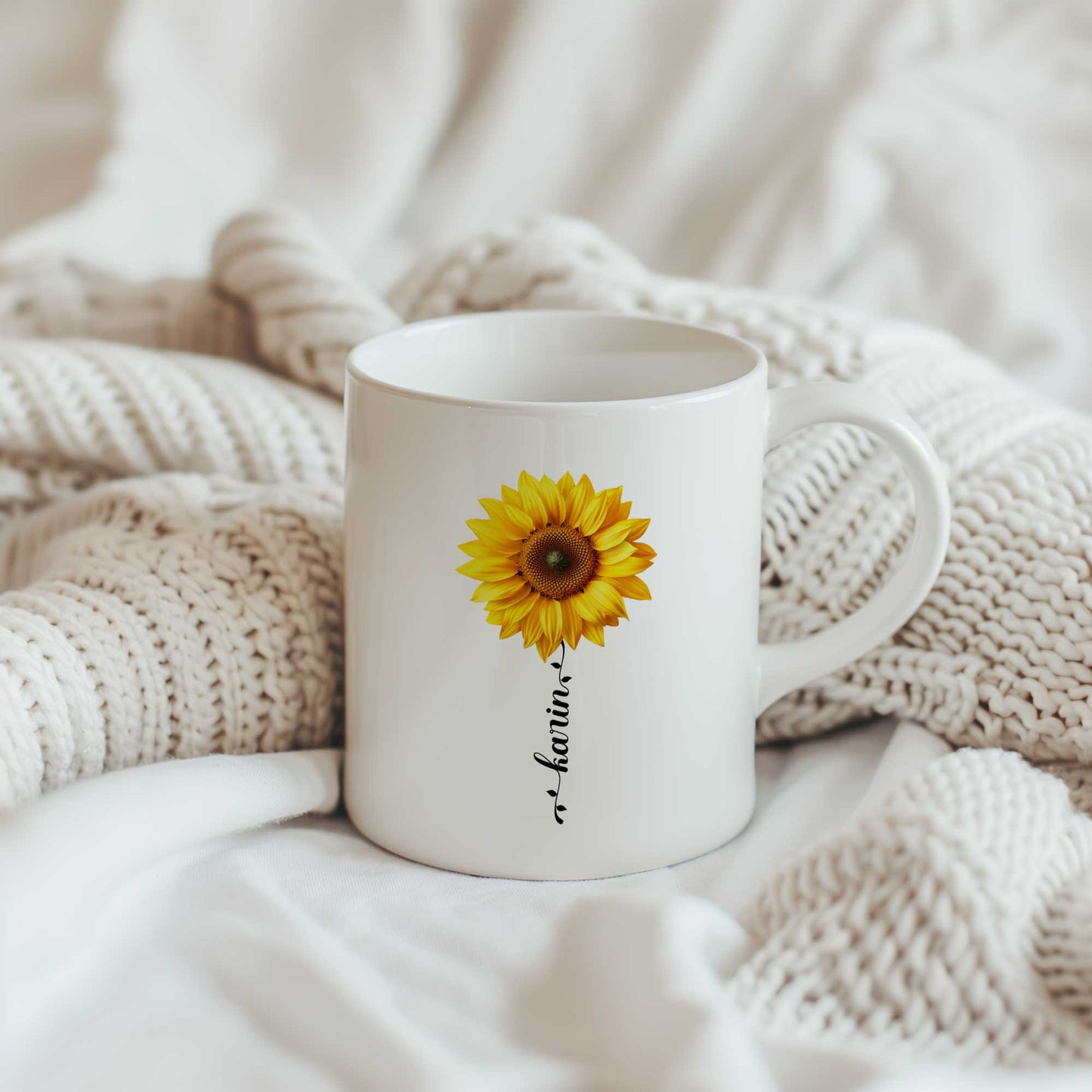 Witte keramische mok met grote gele zonnebloem en gepersonaliseerde naam als bloemsteel, geplaatst op een beige gebreide deken.