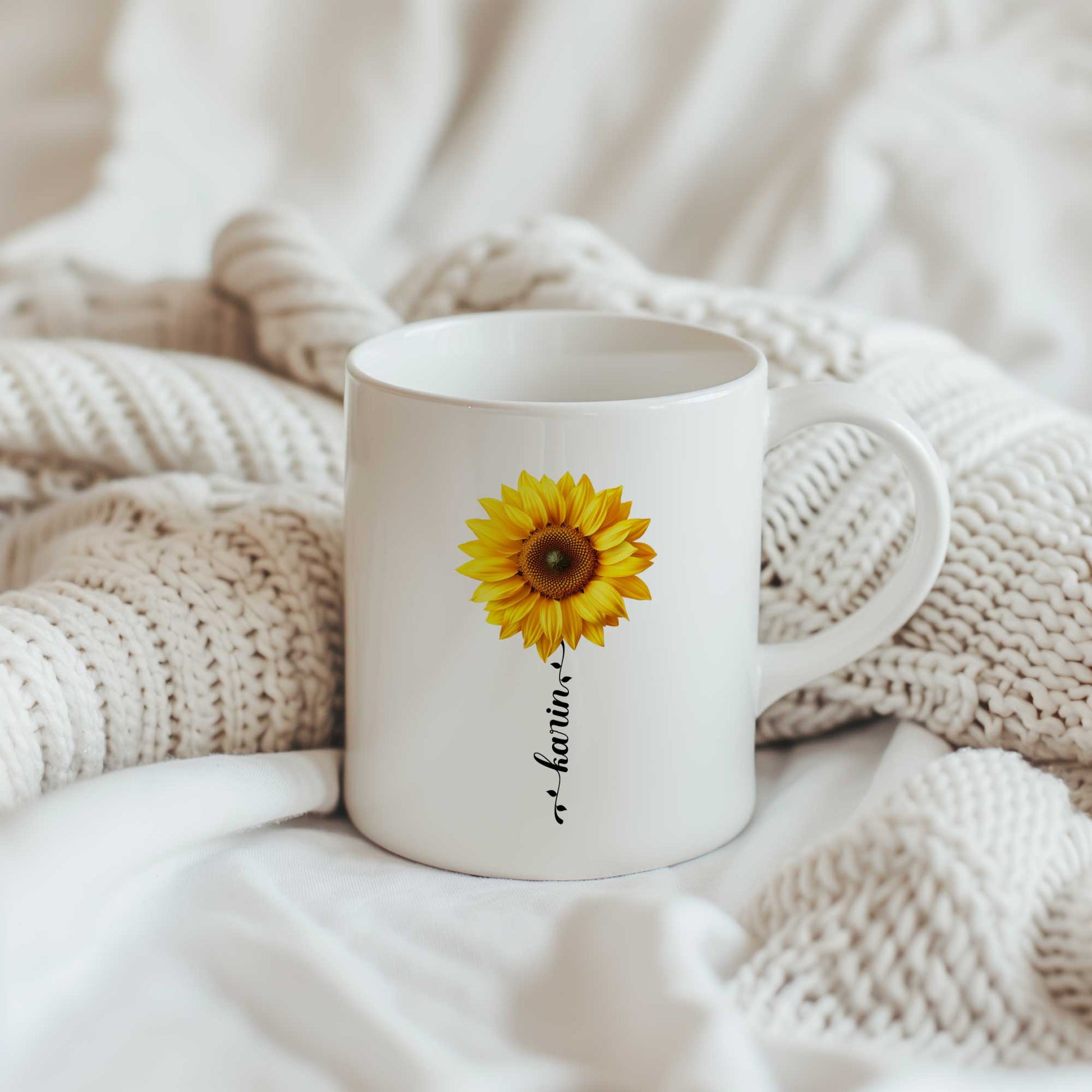 Witte keramische mok met grote gele zonnebloem en gepersonaliseerde naam als bloemsteel, geplaatst op een beige gebreide deken.