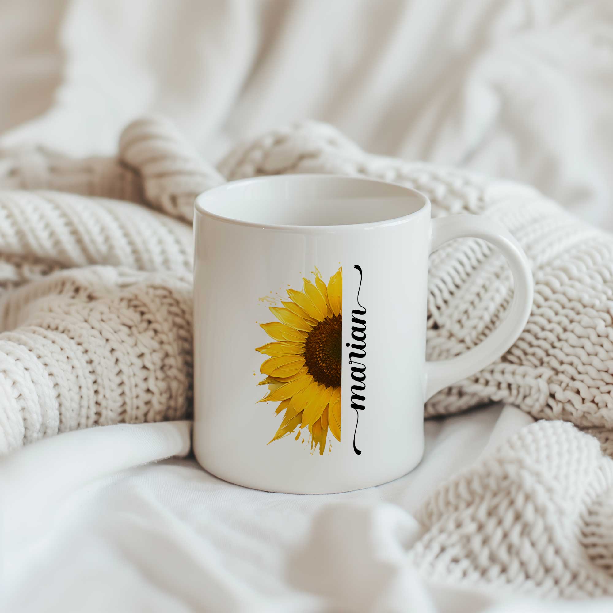 Witte keramische mok met een halve gele zonnebloem en een gepersonaliseerde naam naast de bloem, geplaatst tussen zachte crèmekleurige gebreide dekens.