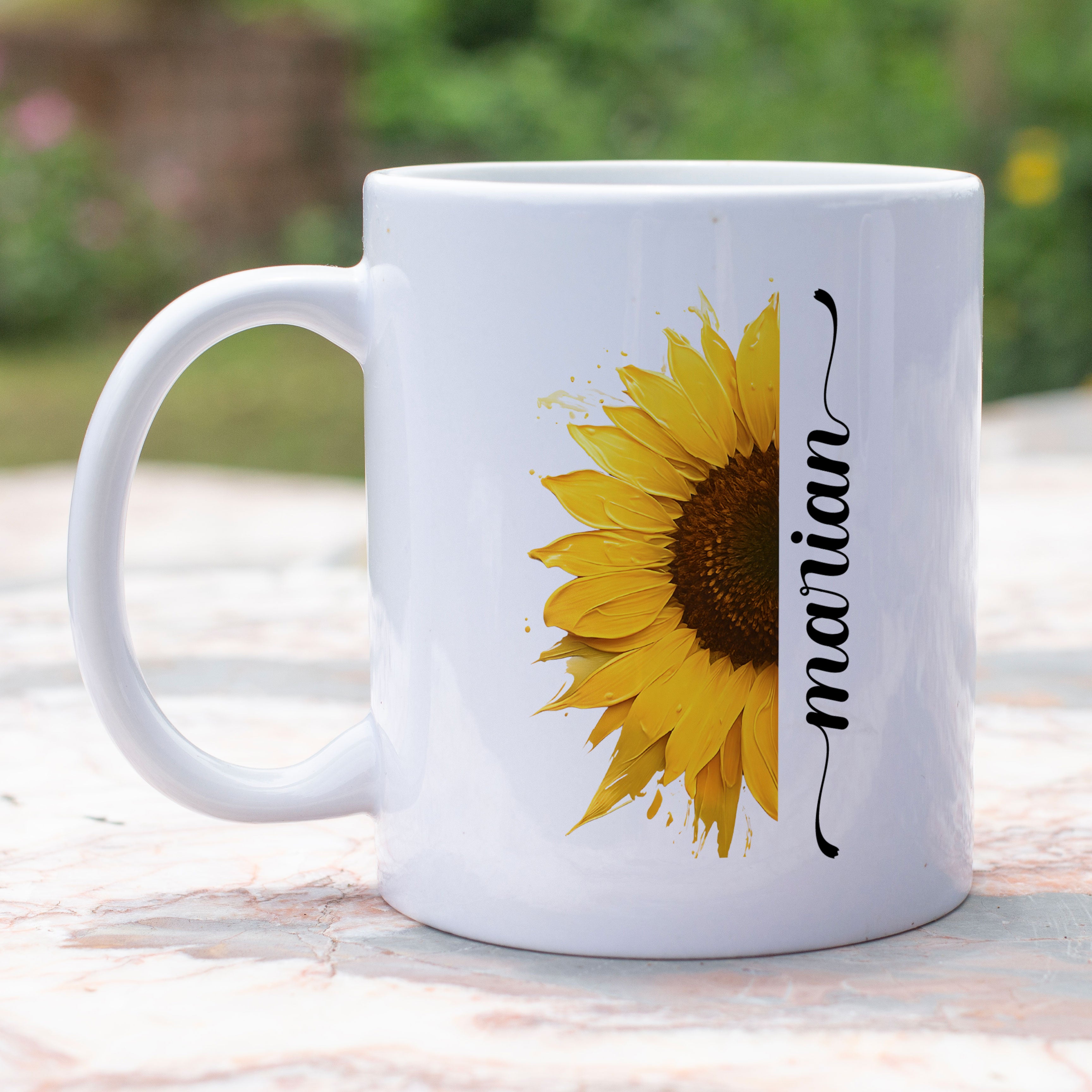 gepersonaliseerde witte mok met halve zonnebloem in acryl verf effect met op de andre helft een sierlijke naam in het zwart buiten in de tuin op een tuintafel