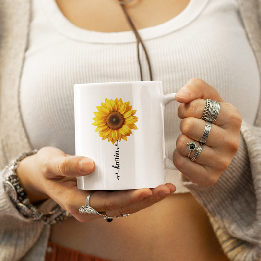 een vrouw met een mok in haar hand het is  een witte mok met zonnebloem en aanpasbare naam in een zwart sierlijke lettertype in de vorm van een bloemstengel