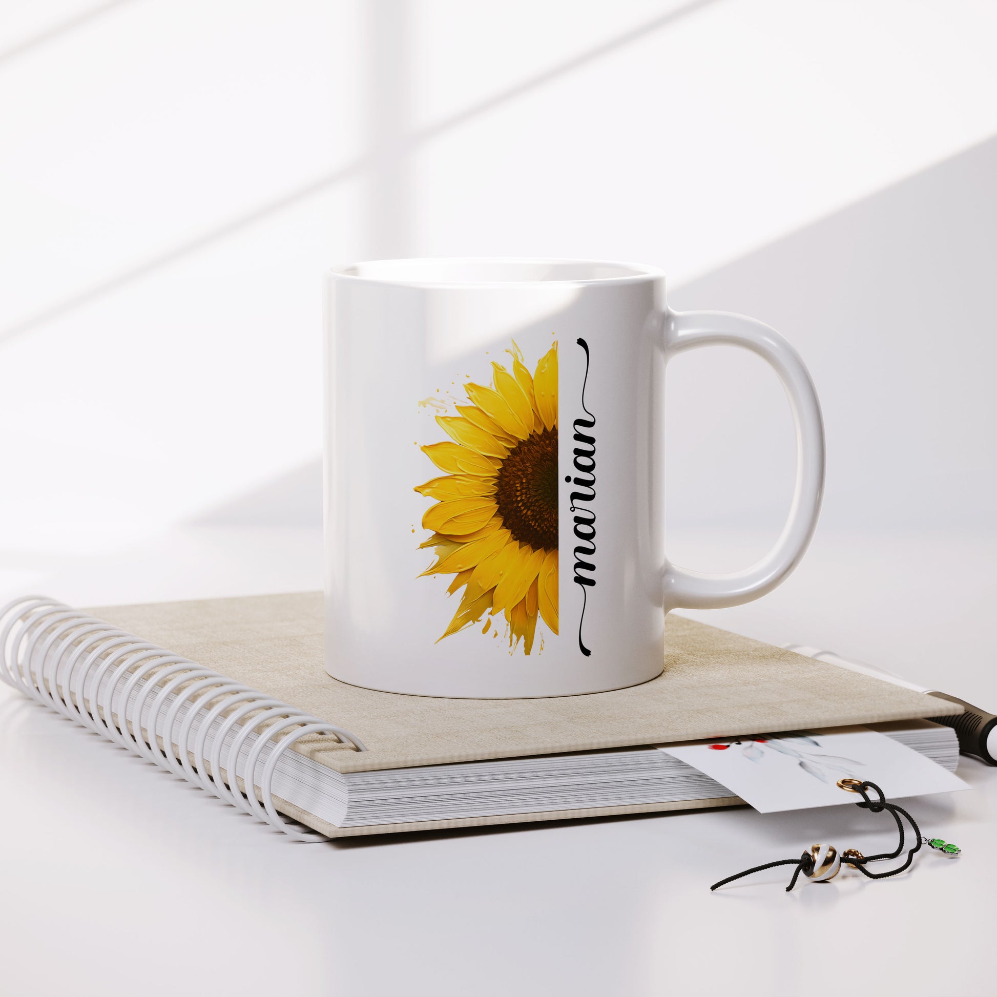 gepersonaliseerde witte mok met halve zonnebloem in acryl verf effect met op de andre helft een sierlijke naam in het zwart op een notitieboek met bladwijzer