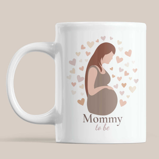 Witte keramische Mommy To Be mok op beige achtergrond met minimalistische illustratie van zwangere vrouw en pastel hartjes.