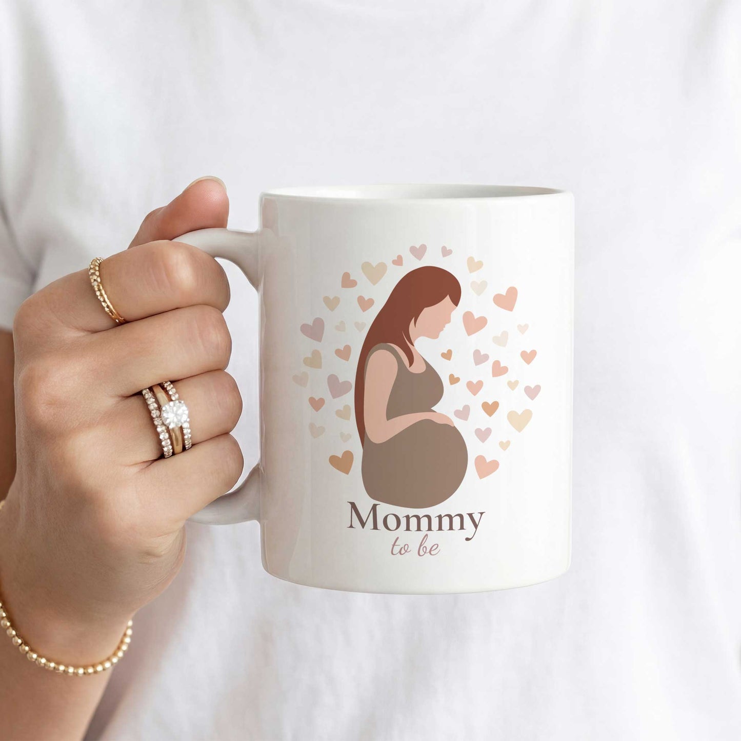 Witte Mommy To Be mok met illustratie van een zwangere vrouw en hartjes, vastgehouden door een vrouw met gouden sieraden en witte kleding.