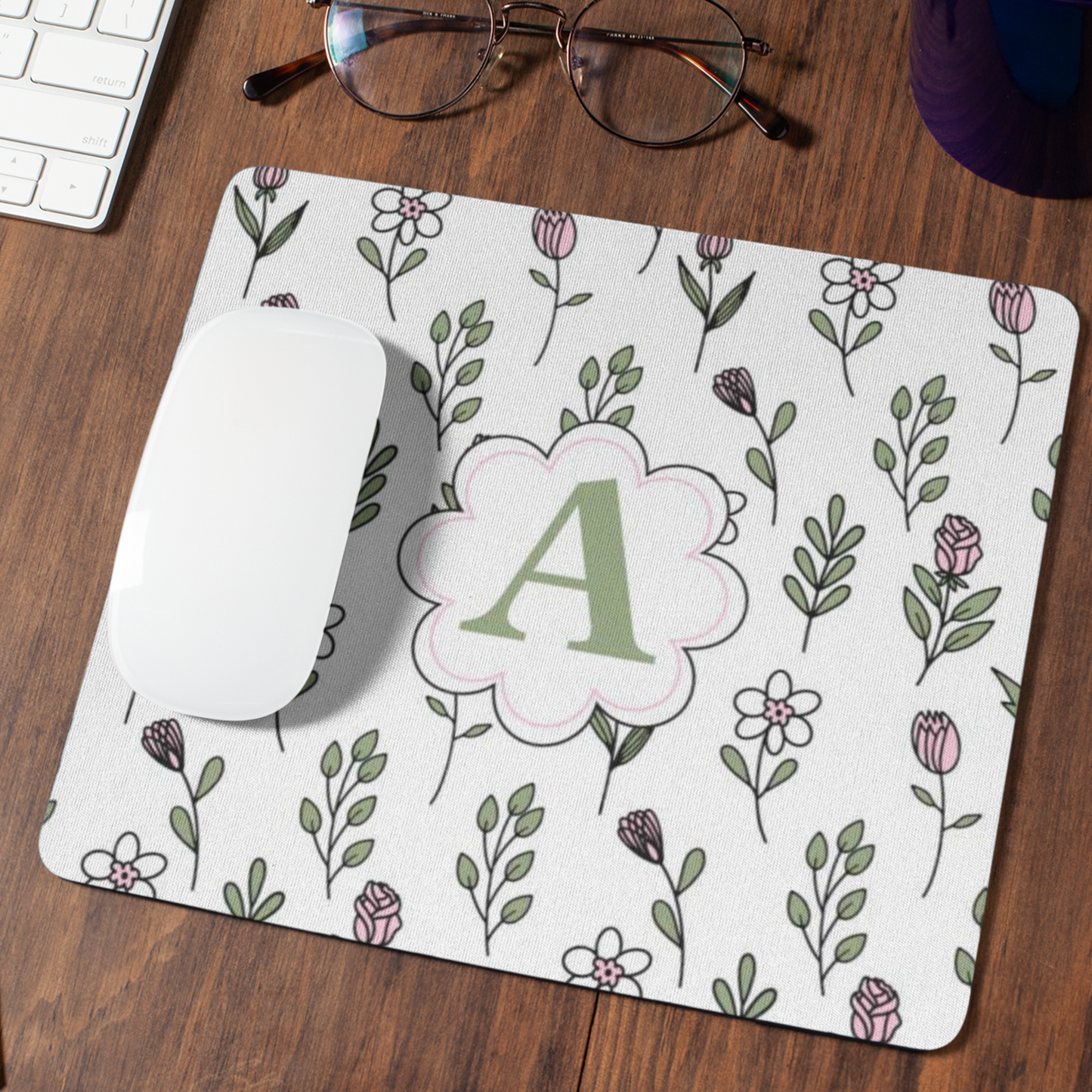Muismat met antislip onderkant, met patroon van lijn bloemen in licht groene en roze kleuren en in het midden een Monogram van de letter van iemands naam die aanpasbaar is liggend op een donker houten bureau met een lessbril en een witte muis