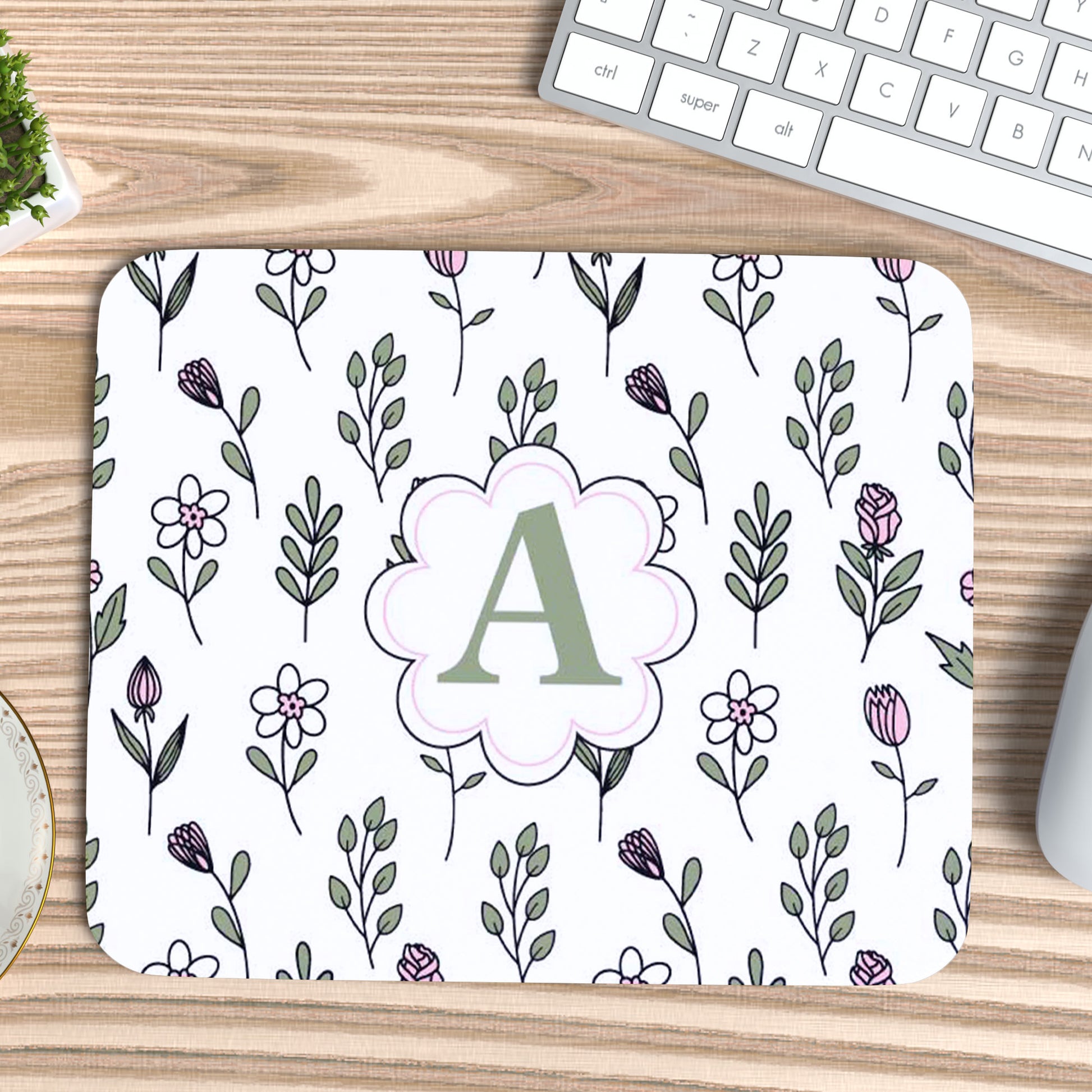 Muismat met antislip onderkant, met patroon van lijn bloemen in licht groene en roze kleuren en in het midden een Monogram van de letter van iemands naam die aanpasbaar is naast een wit oetsenbord op een licht houten bureau