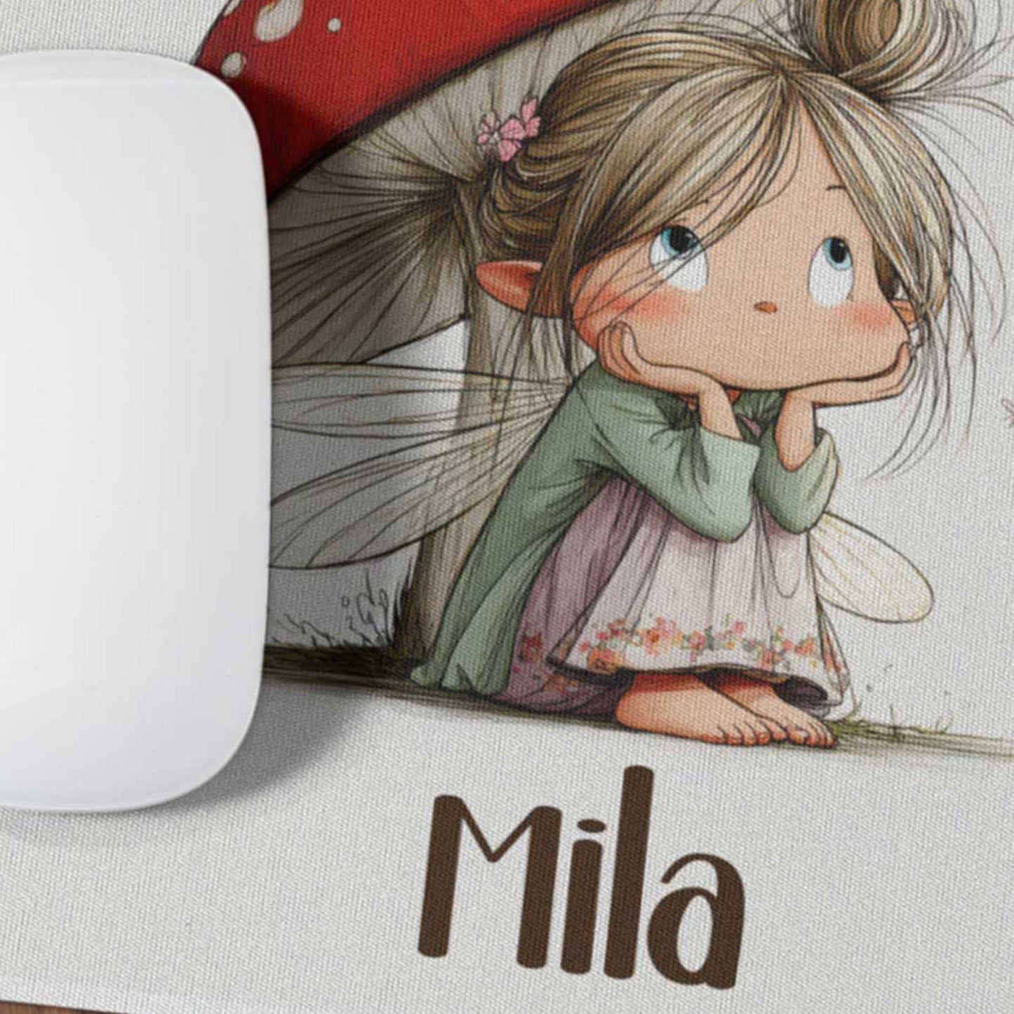 Close-up van een muismat met een illustratie van een fee onder een rode paddenstoel en de naam Mia naast een witte computermuis.