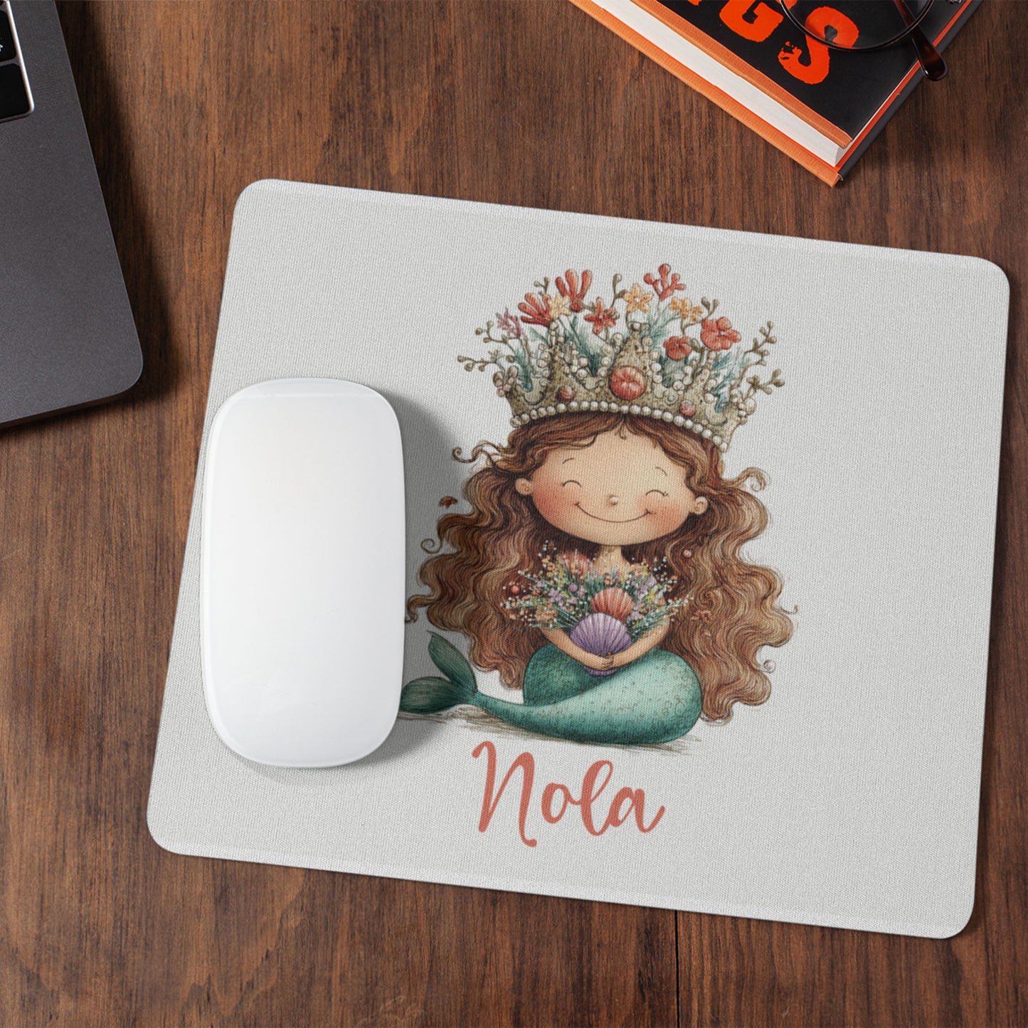 Witte muismat met een zeemeermin met koraalkroon en de naam Nola op een houten bureau naast een witte computermuis, laptop en boek.