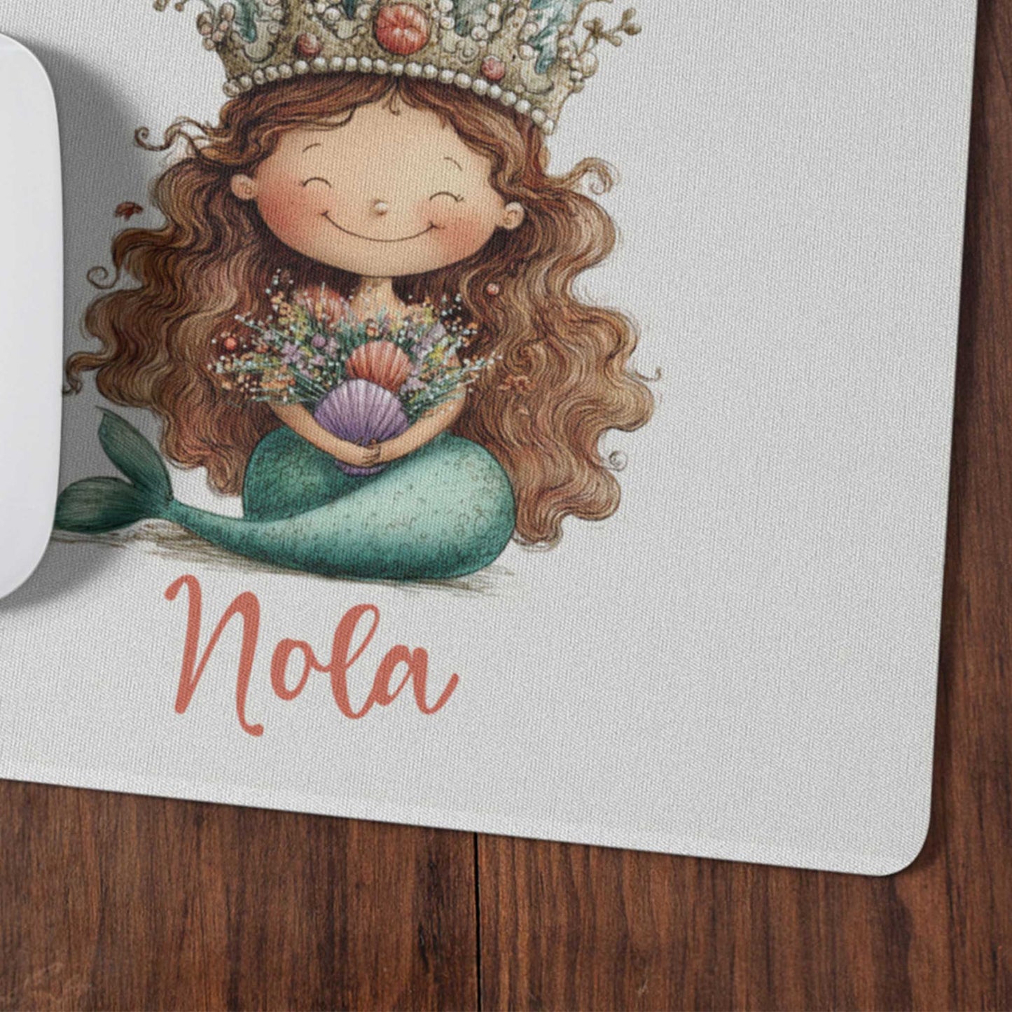 Close-up van een witte muismat met een zeemeermin met lange bruine haren, een koraalkroon en de naam Nola op een houten bureau.